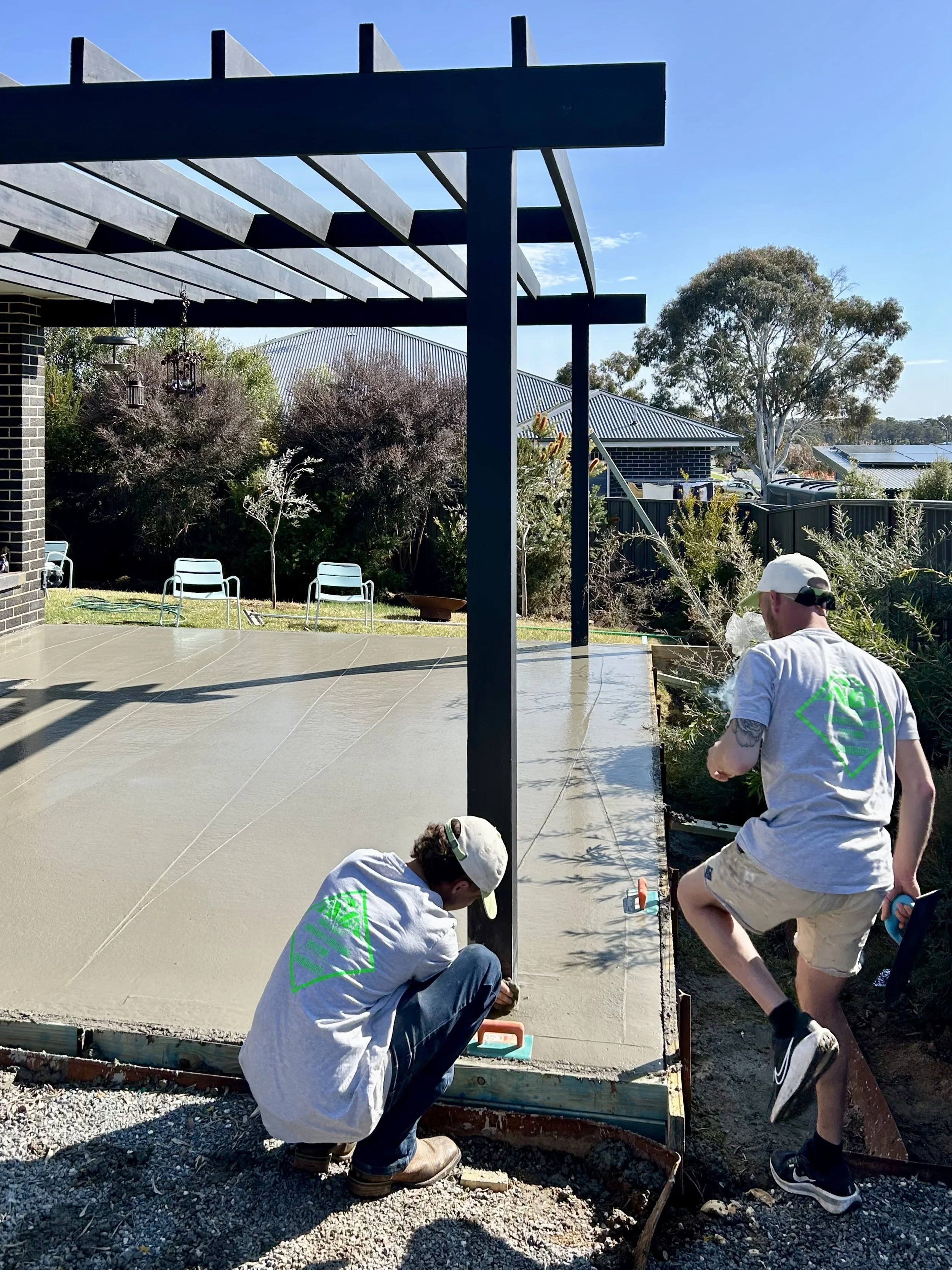 Two workers in casual clothes and white hats are working on a concrete patio, with one crouching and measuring and the other standing and preparing to pour or finish the concrete. The patio has a black metal pergola overhead and is bordered by a gard