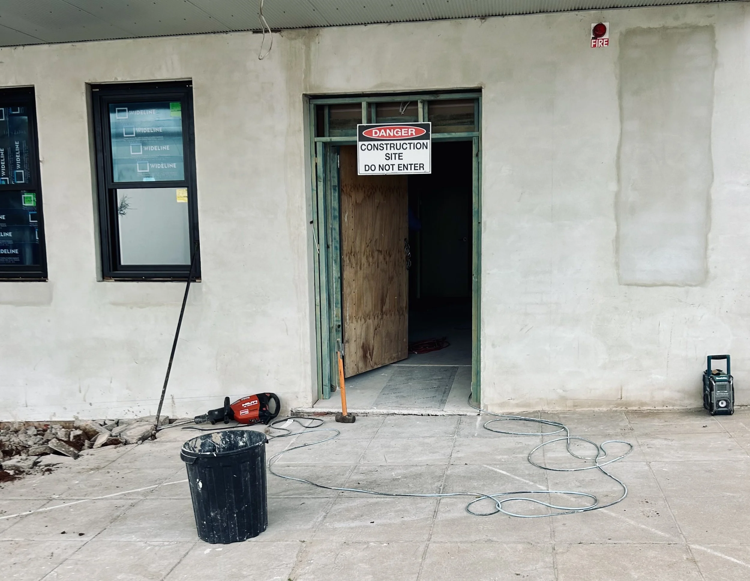 Construction site with a partially open doorway marked 'Danger Construction Site Do Not Enter', windows, a black trash can, construction tools, and cables on the ground.