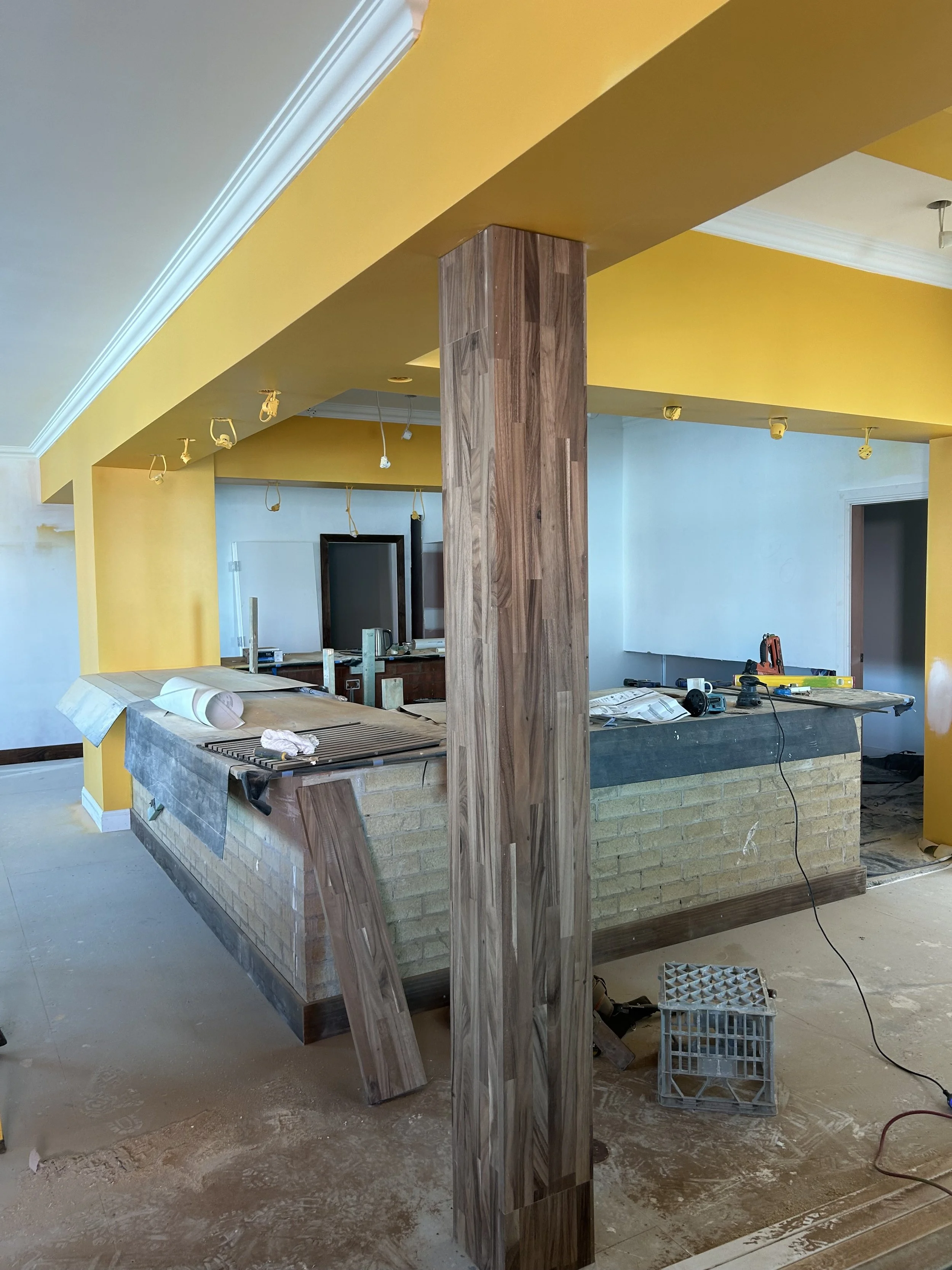 Kitchen under construction with yellow walls, wooden column, and brick counter, tools and construction materials scattered around.