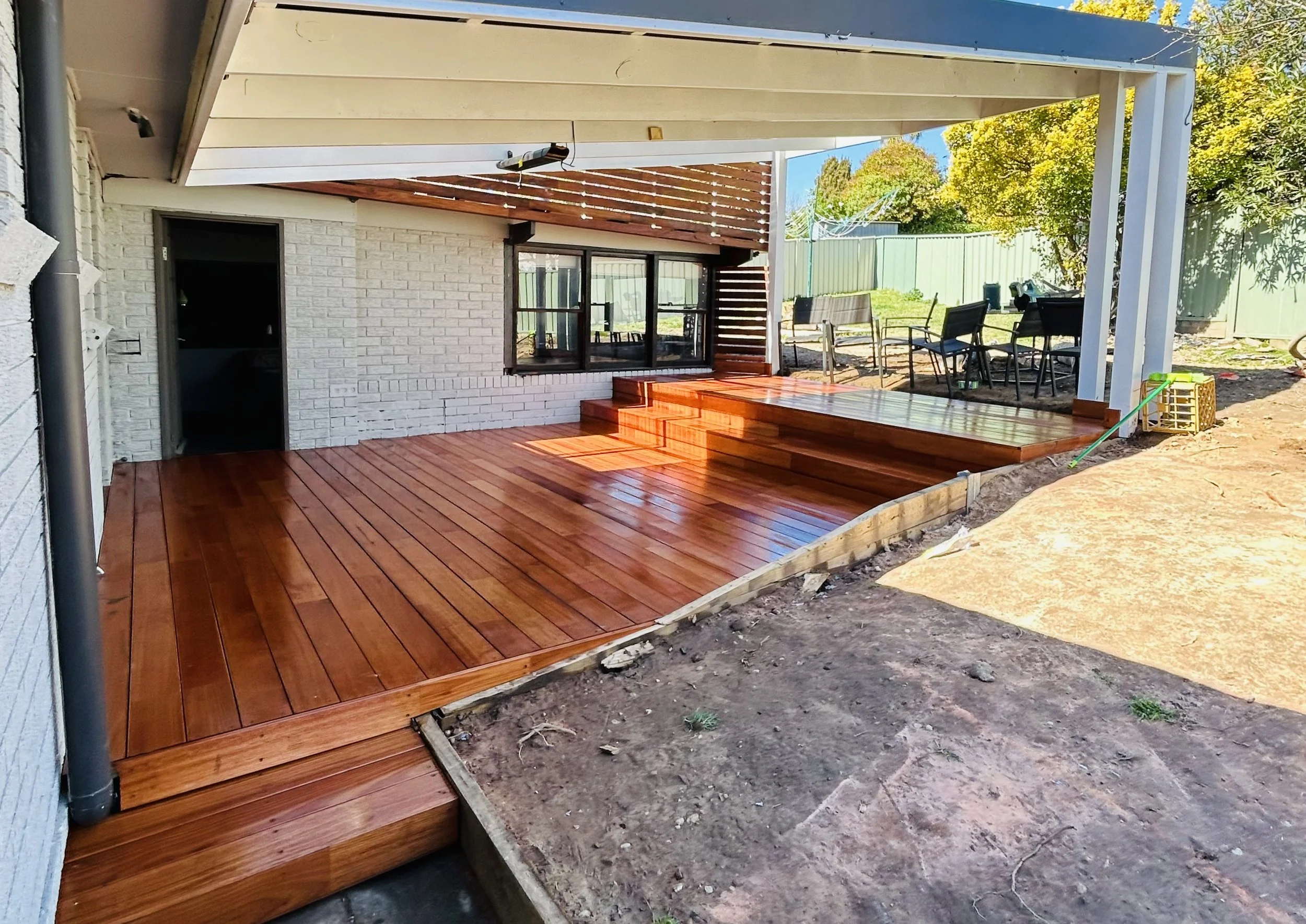 Finished wooden deck with stairs on backyard patio, surrounded by outdoor seating and a green fence, with trees in the background.