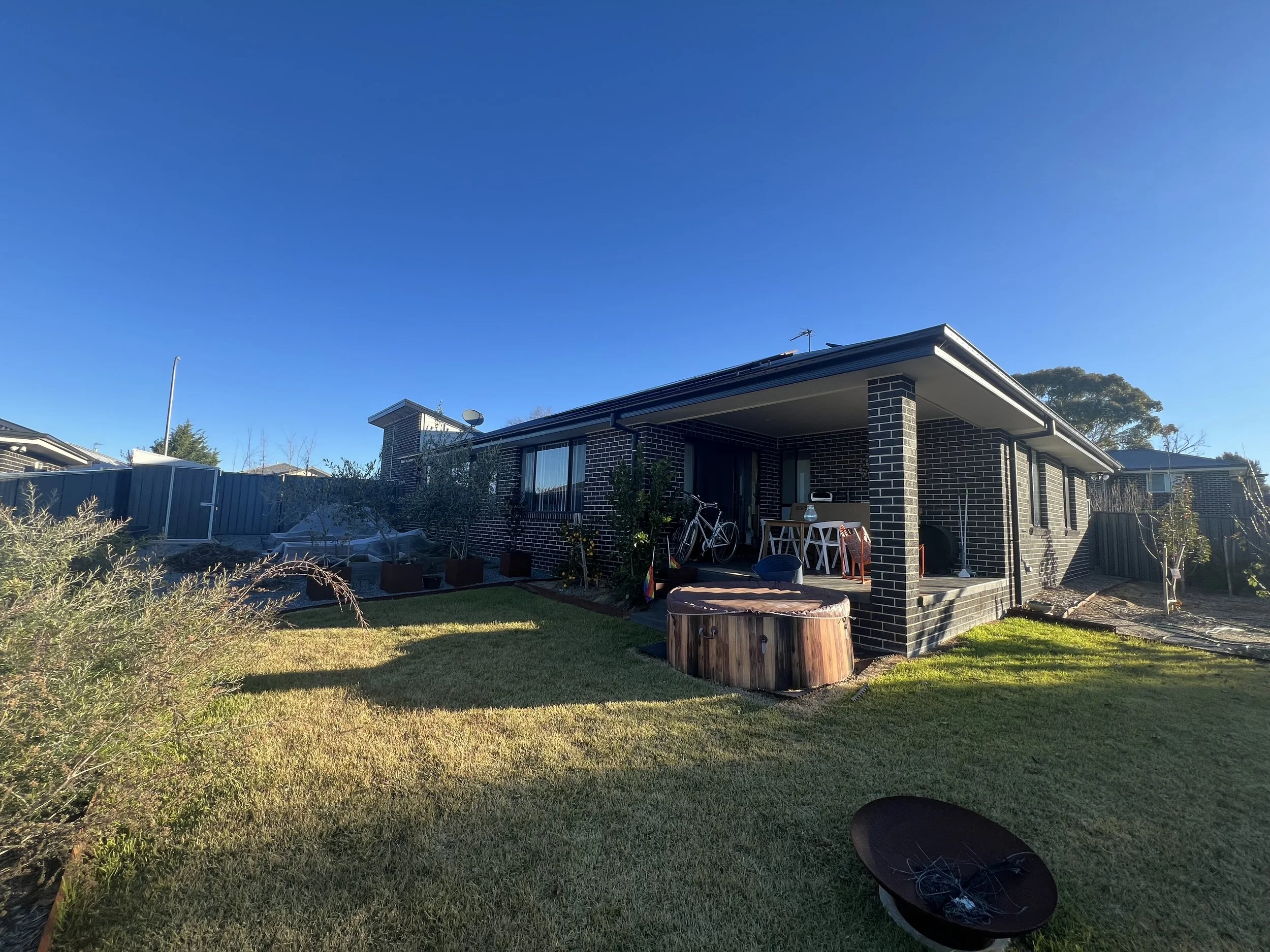 Backyard view of a brick house with a covered patio, outdoor furniture, bicycles, a hot tub, laundry, and a lawn under a clear blue sky.