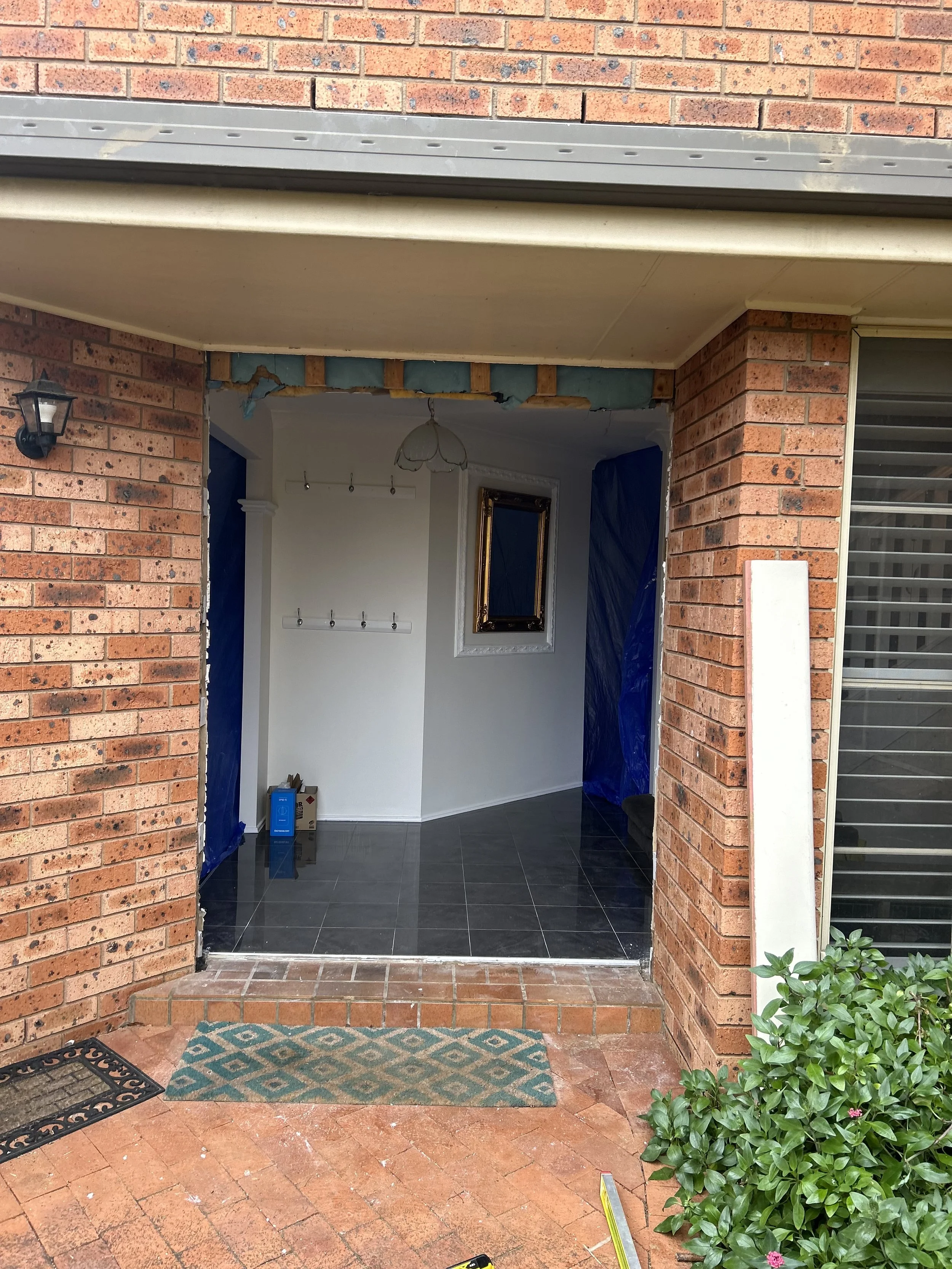 Porch entrance of house under construction or renovation, with brick walls, a doormat, small step, and interior glimpse showing tile flooring, a white wall, and a window, covered partially with blue protective plastic.
