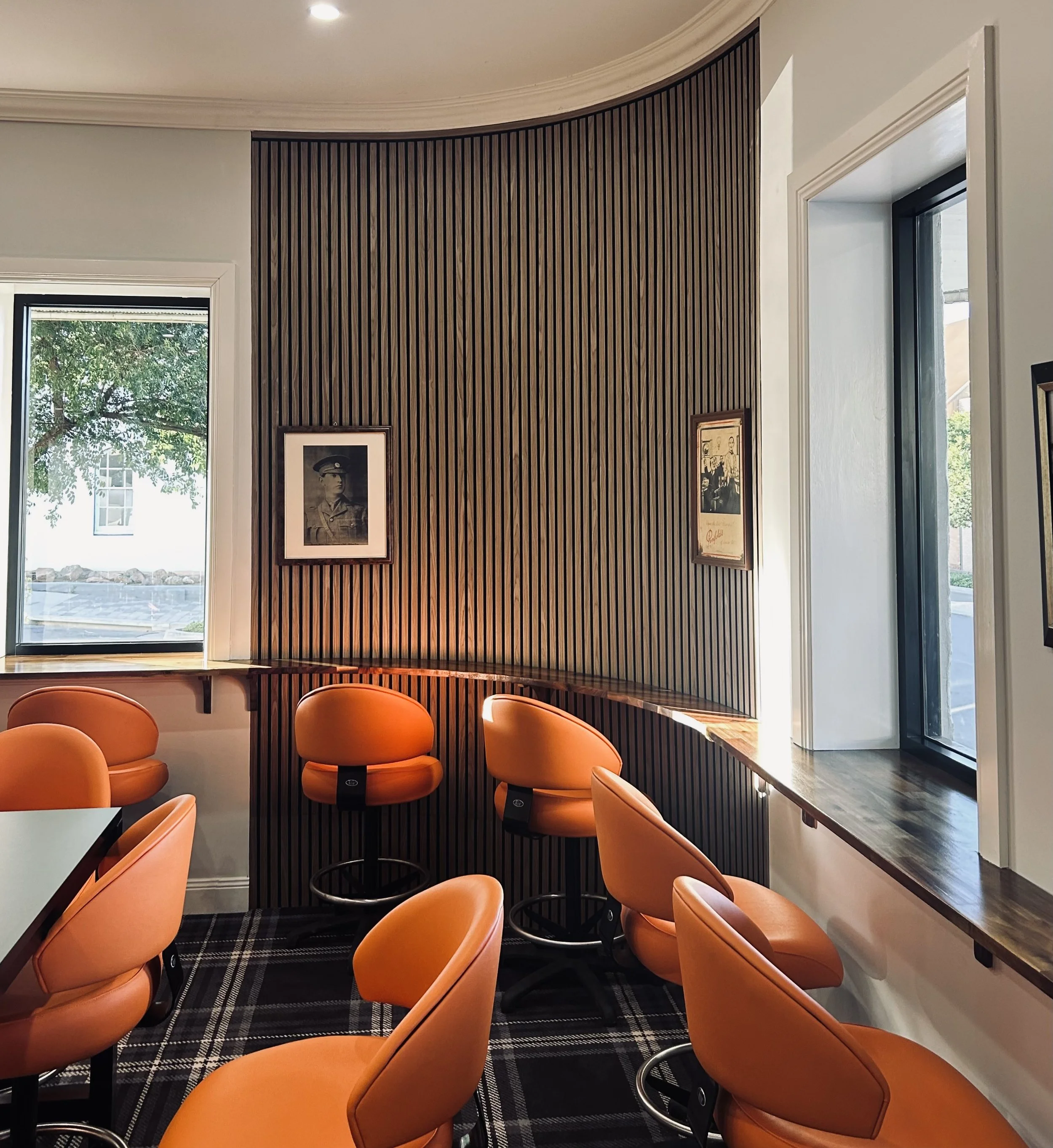 A cafe or restaurant interior with orange chairs, a curved wooden windowsill, a dark wood slat accent wall, and framed black and white photographs. Large windows allow natural light to brighten the space.