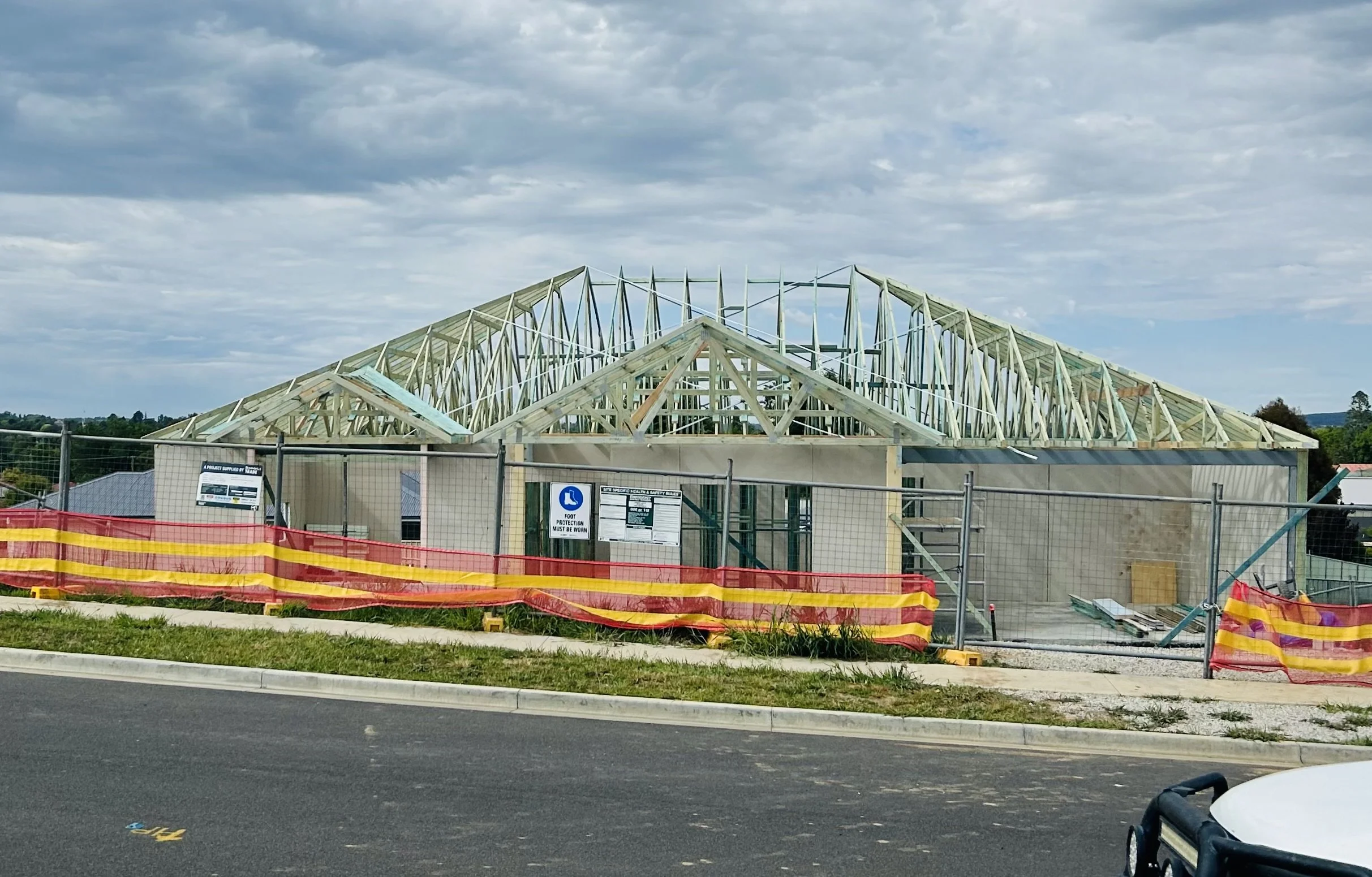 A house under construction with a visible roof frame, surrounded by a construction fence with warning flags and signs, and a cloudy sky overhead.