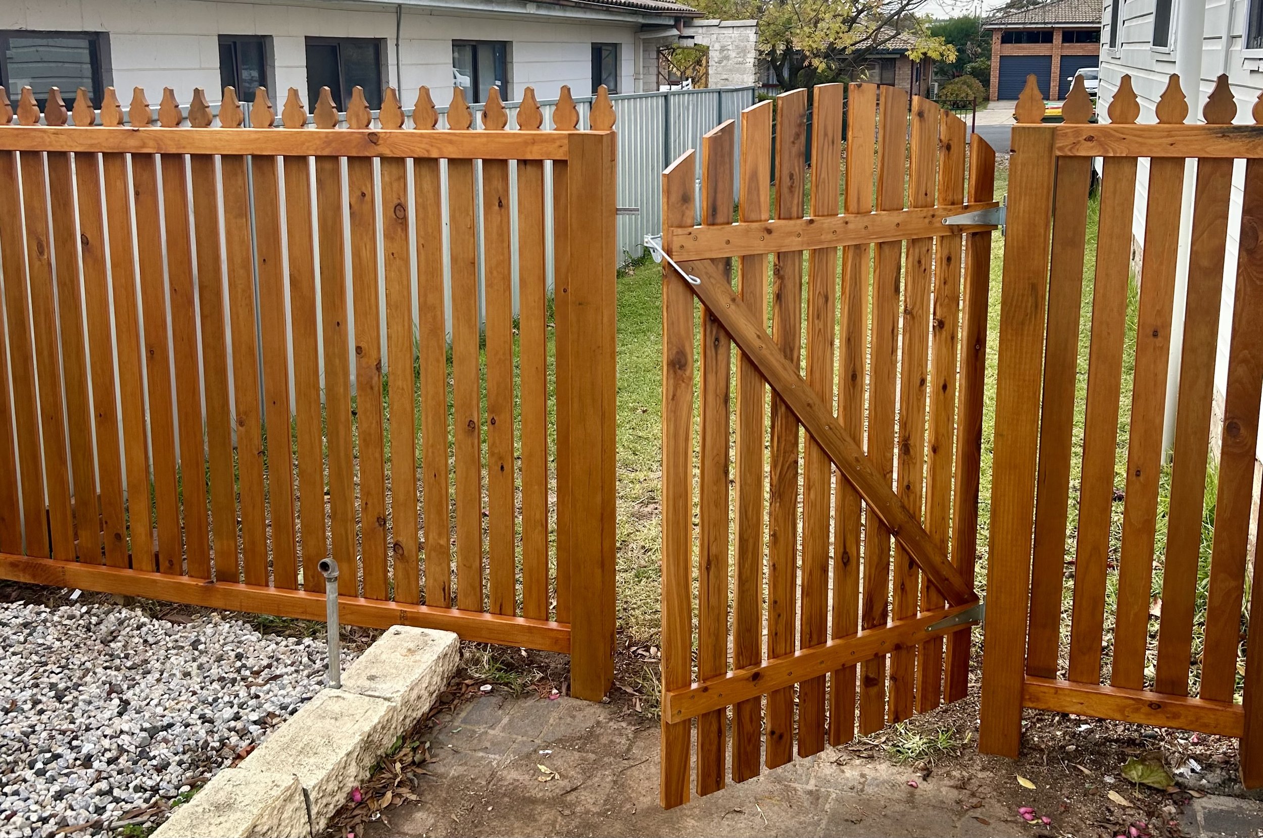 A wooden fence with a gate, installed on a grassy backyard, with gravel and paving stones at the base, and neighboring houses visible in the background.