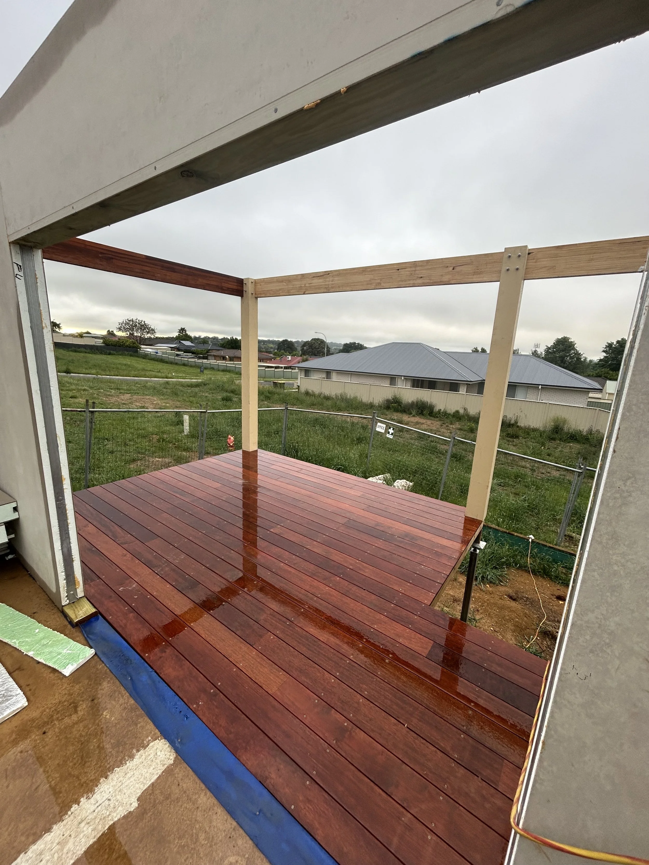 Partially constructed outdoor patio with newly installed wooden decking, supported by vertical posts, with overcast sky and surrounding grassy area and houses in the background.