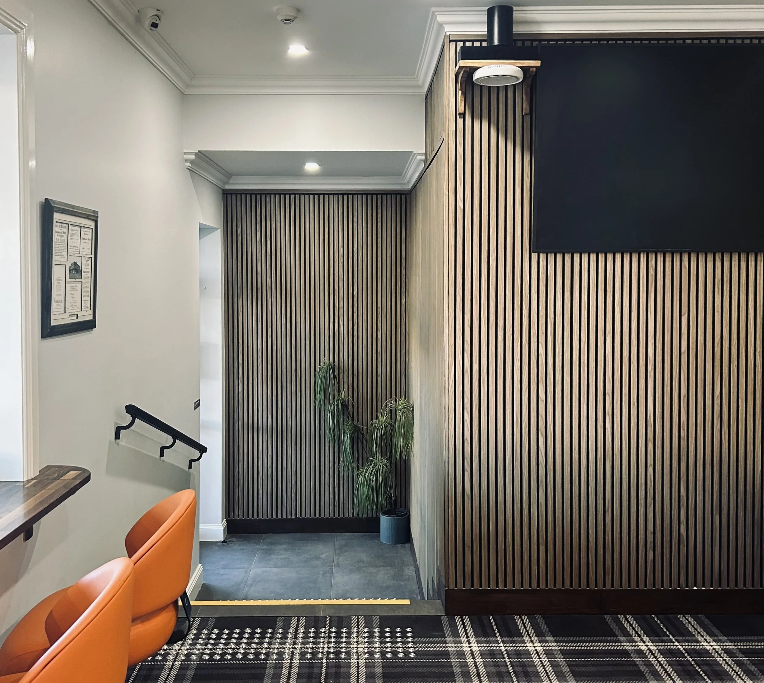 Interior of a modern pub area with white walls, wooden slatted wall panels, a potted plant, orange chairs, and a textured black and beige carpet.