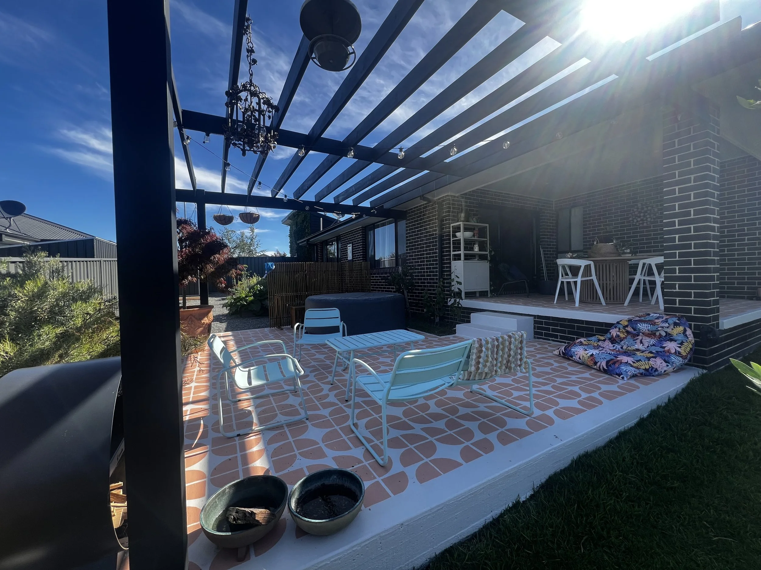 Outdoor patio with modern furniture, hanging lights, and potted plants, attached to a brick house under a sunny, blue sky.
