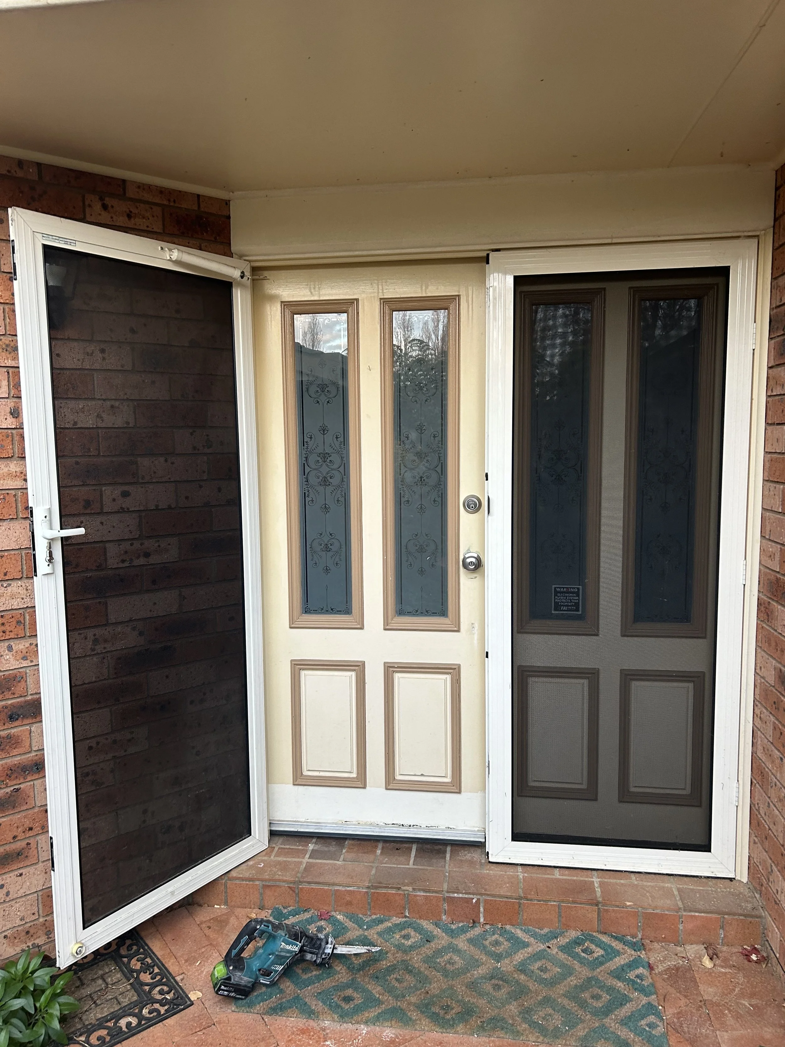 Front porch with three screens: left screen is black, middle door is beige with decorative glass panels, right screen is dark brown with decorative glass panels. A cordless drill and a doormat with a geometric pattern are on the brick porch.
