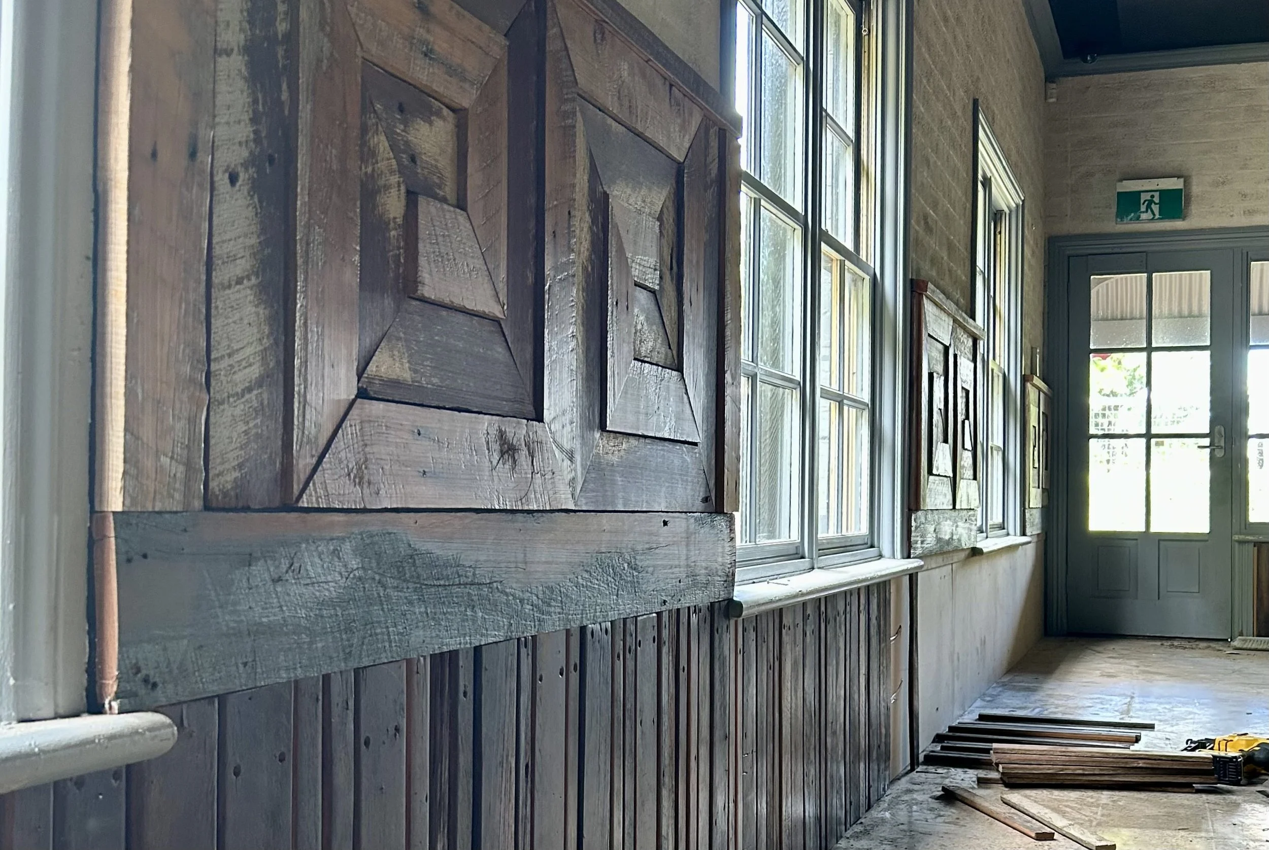 Interior of a building under renovation, with wooden wall panels, abutting large windows, and a door at the end with an emergency exit sign above.