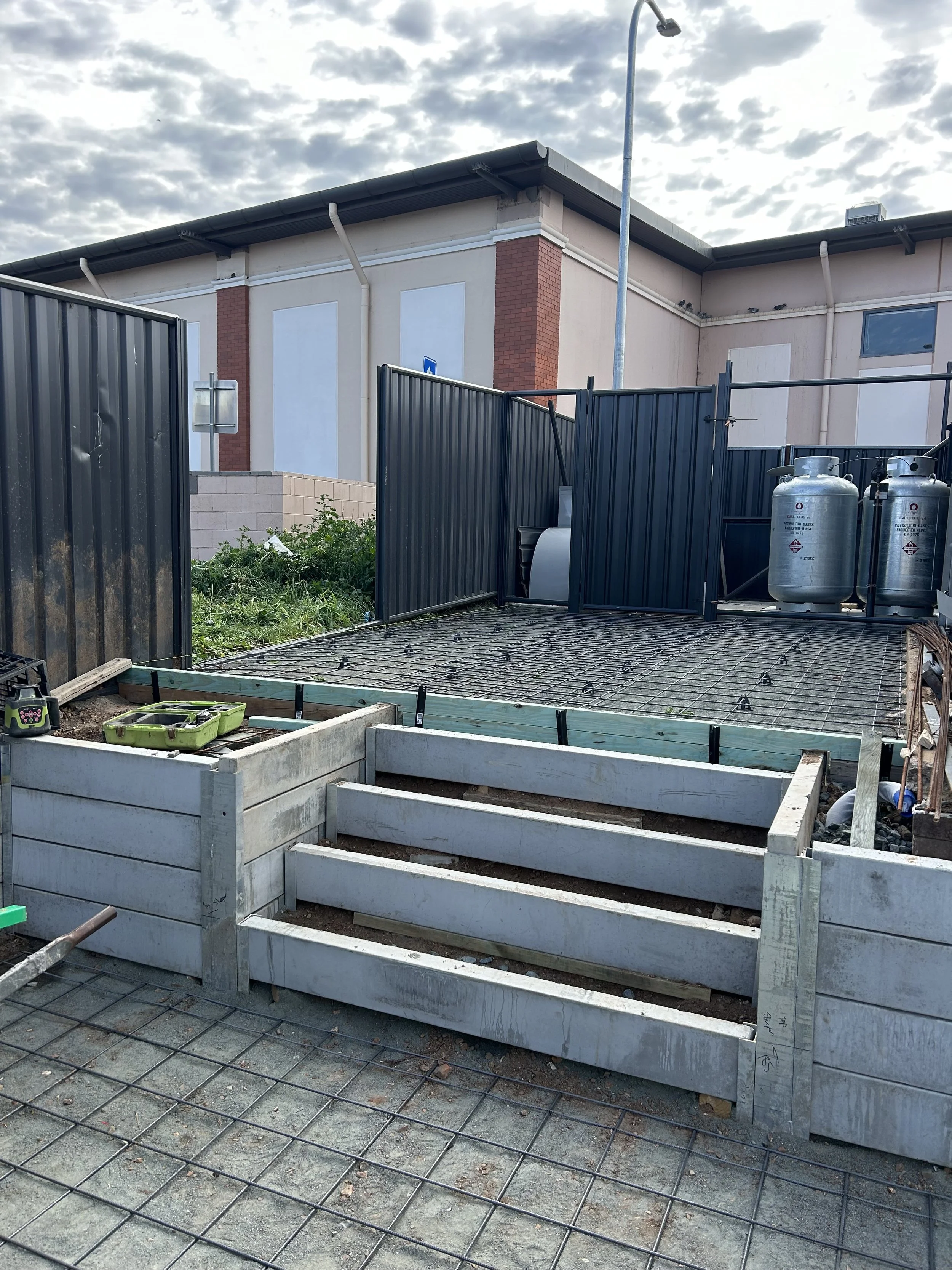 Construction site with wooden stairs leading to a fenced area, concrete blocks, construction tools, and building materials.