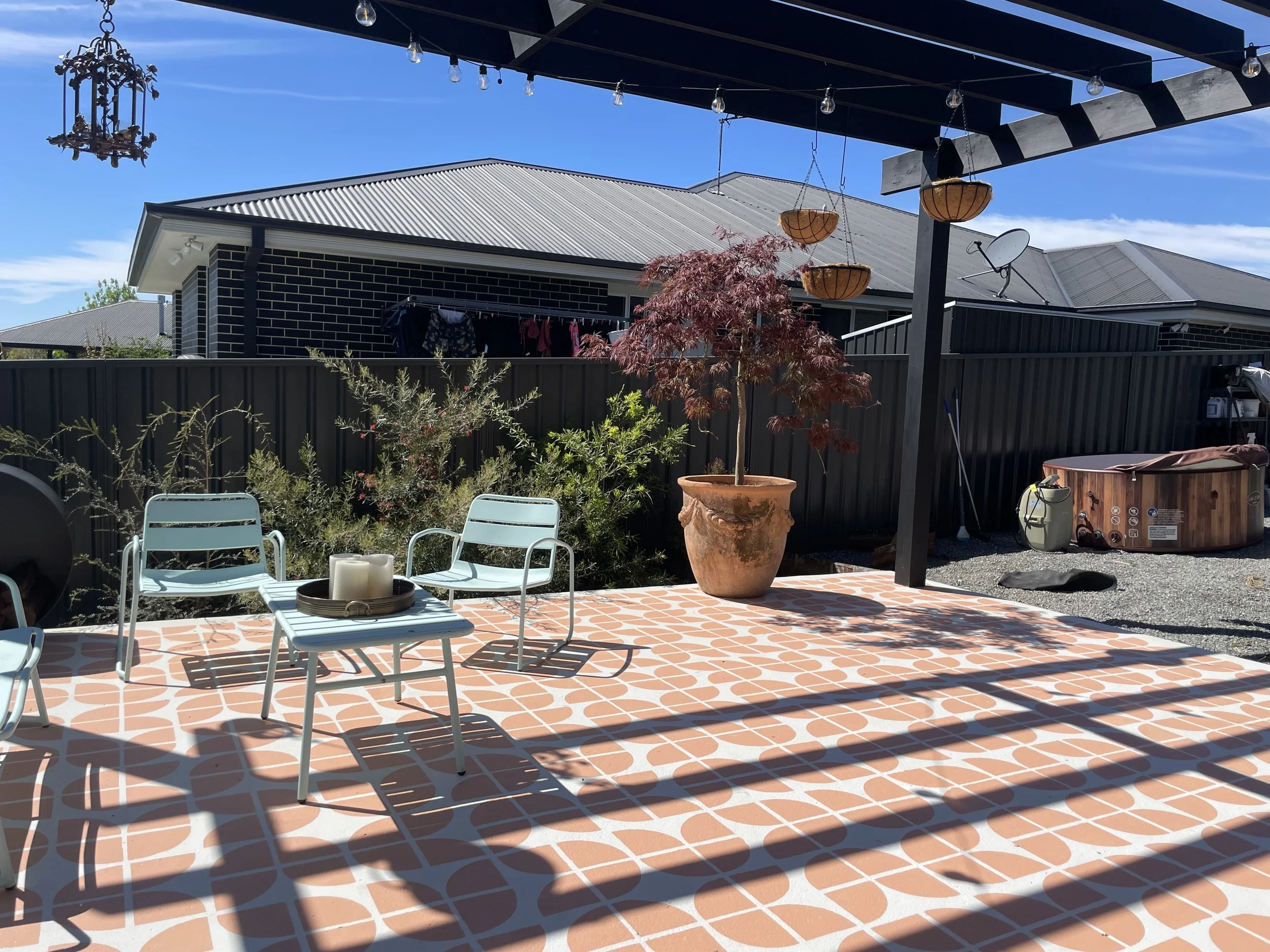 Outdoor patio with patterned tile floor, patio chairs, potted plant, and hanging baskets, under a pergola on a sunny day.