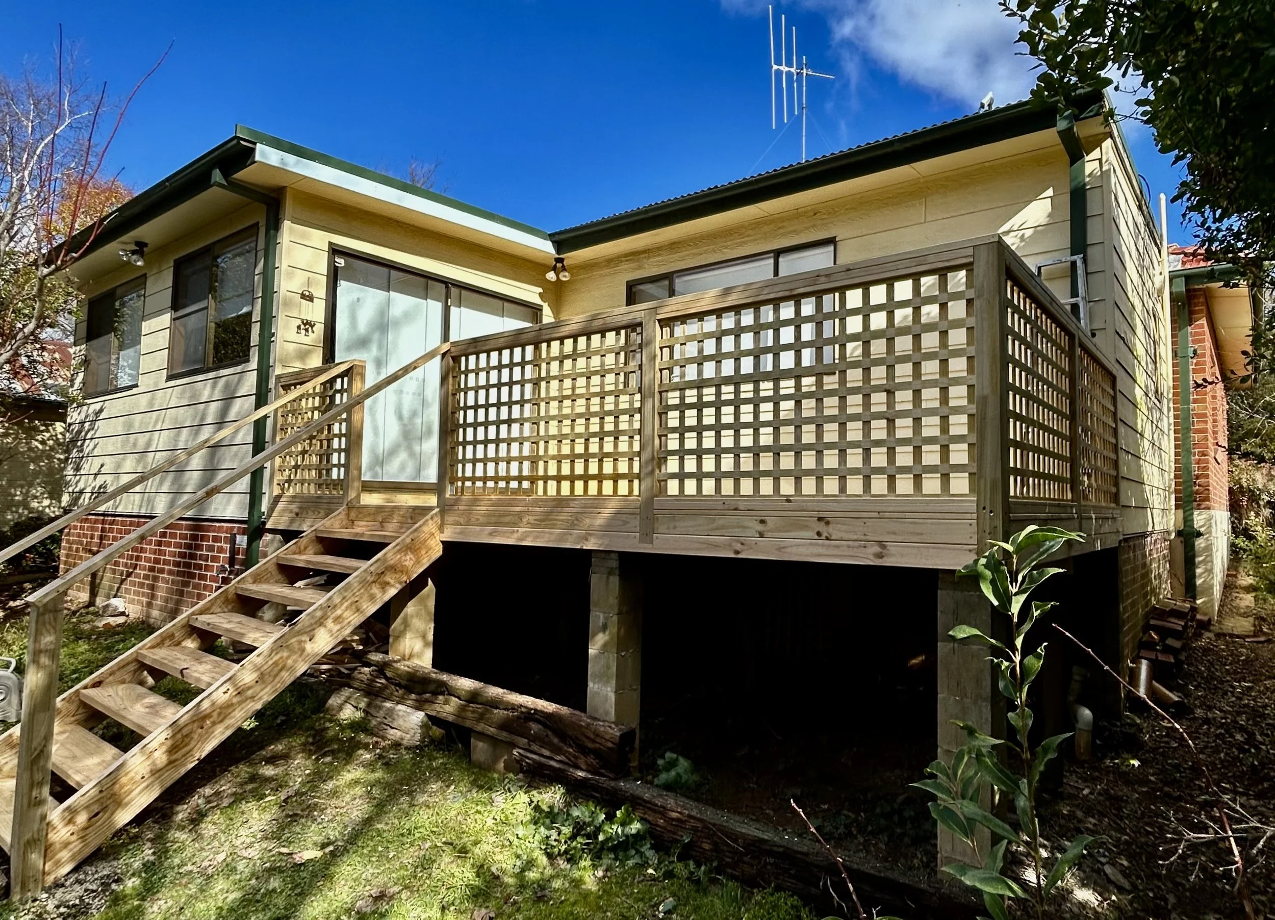 Raised wooden deck with stairs attached to a yellow house with large windows, green trim, and a black roof. The deck is supported by concrete pillars and has lattice fencing. A small plant is visible in the foreground, and the sky is bright blue with