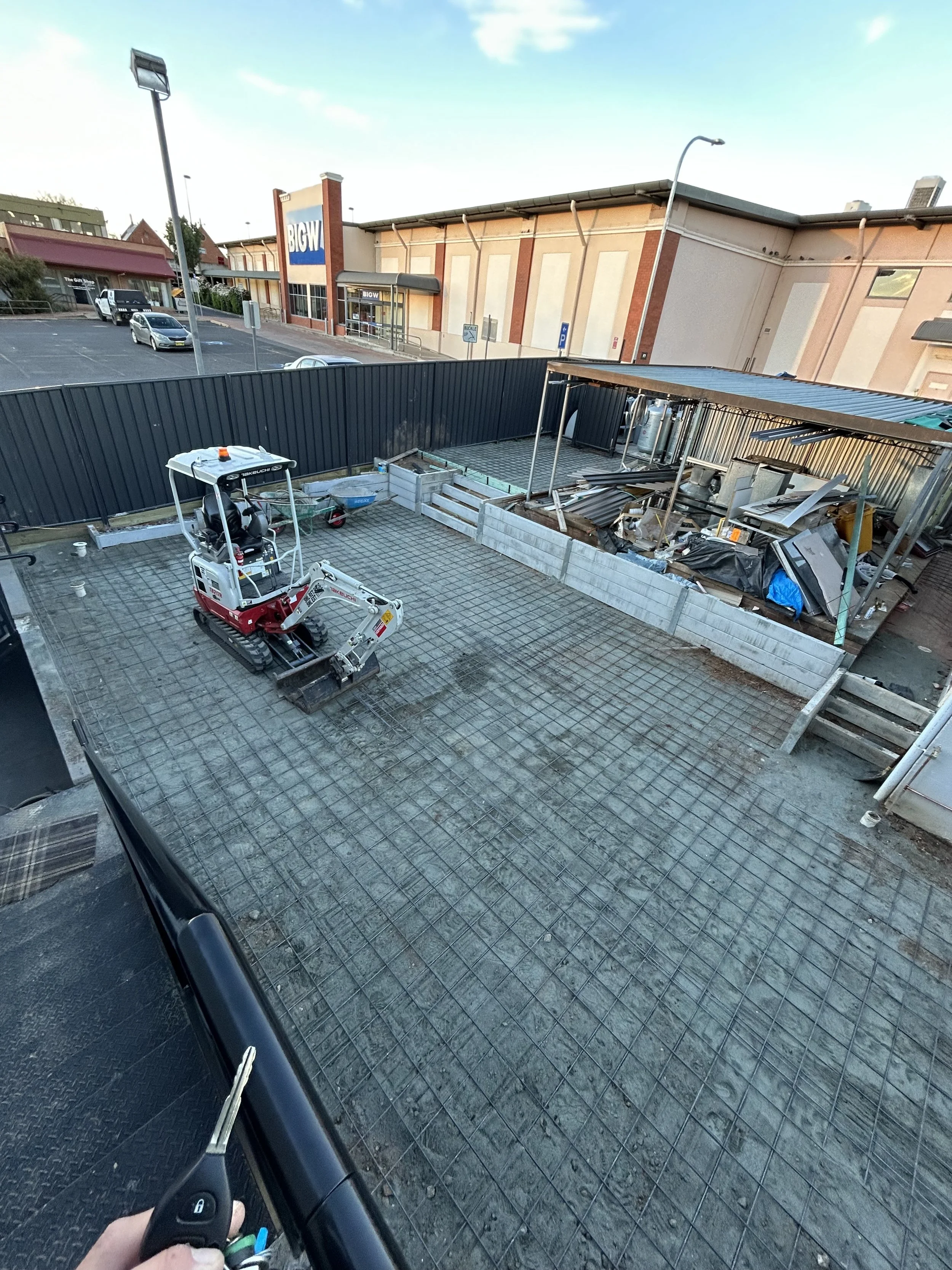 Empty construction site with a small bulldozer and metal rebar grid on the ground, surrounded by a black fence, in front of a retail store called BIG W.