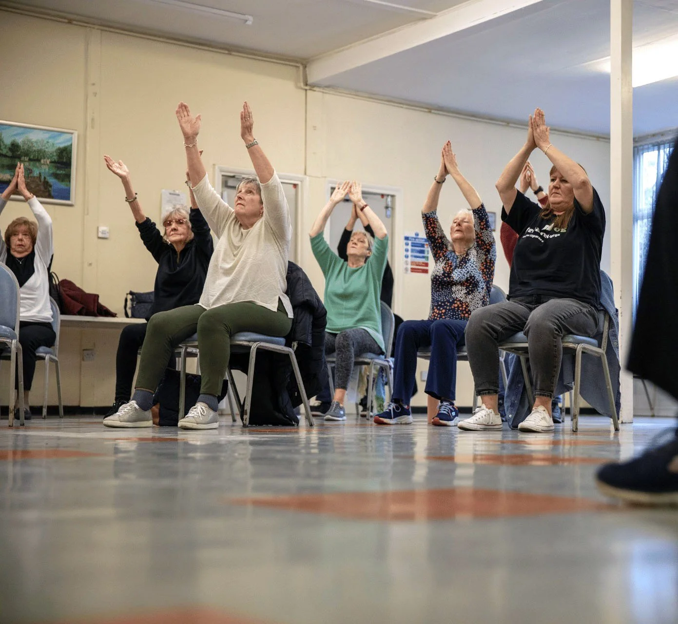Older adults taking part in an Upward Frog chair yoga class, lifting their arms during a seated movement.