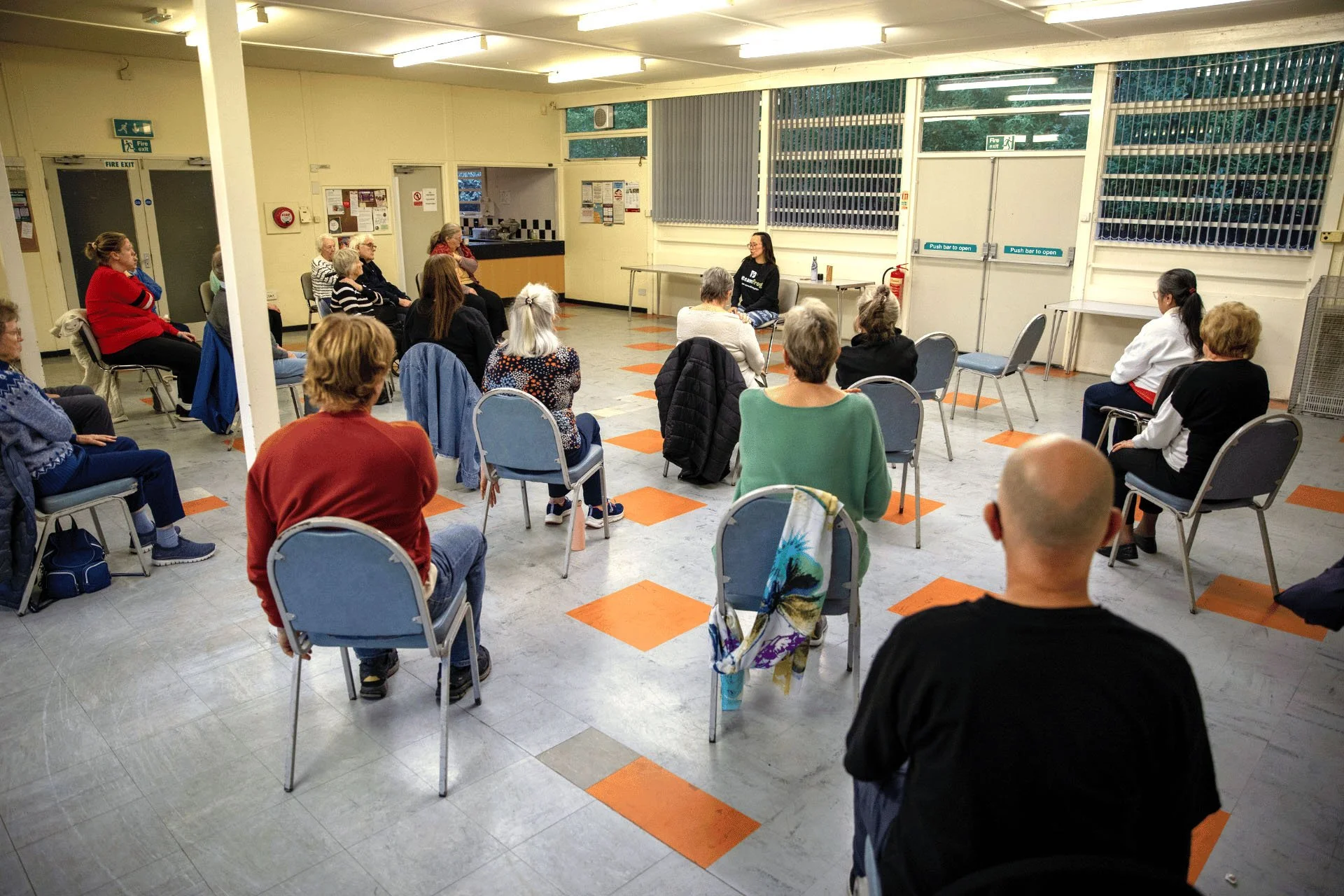 Group of adults taking part in a chair yoga class in a community hall, seated in a circle listening to their instructor.