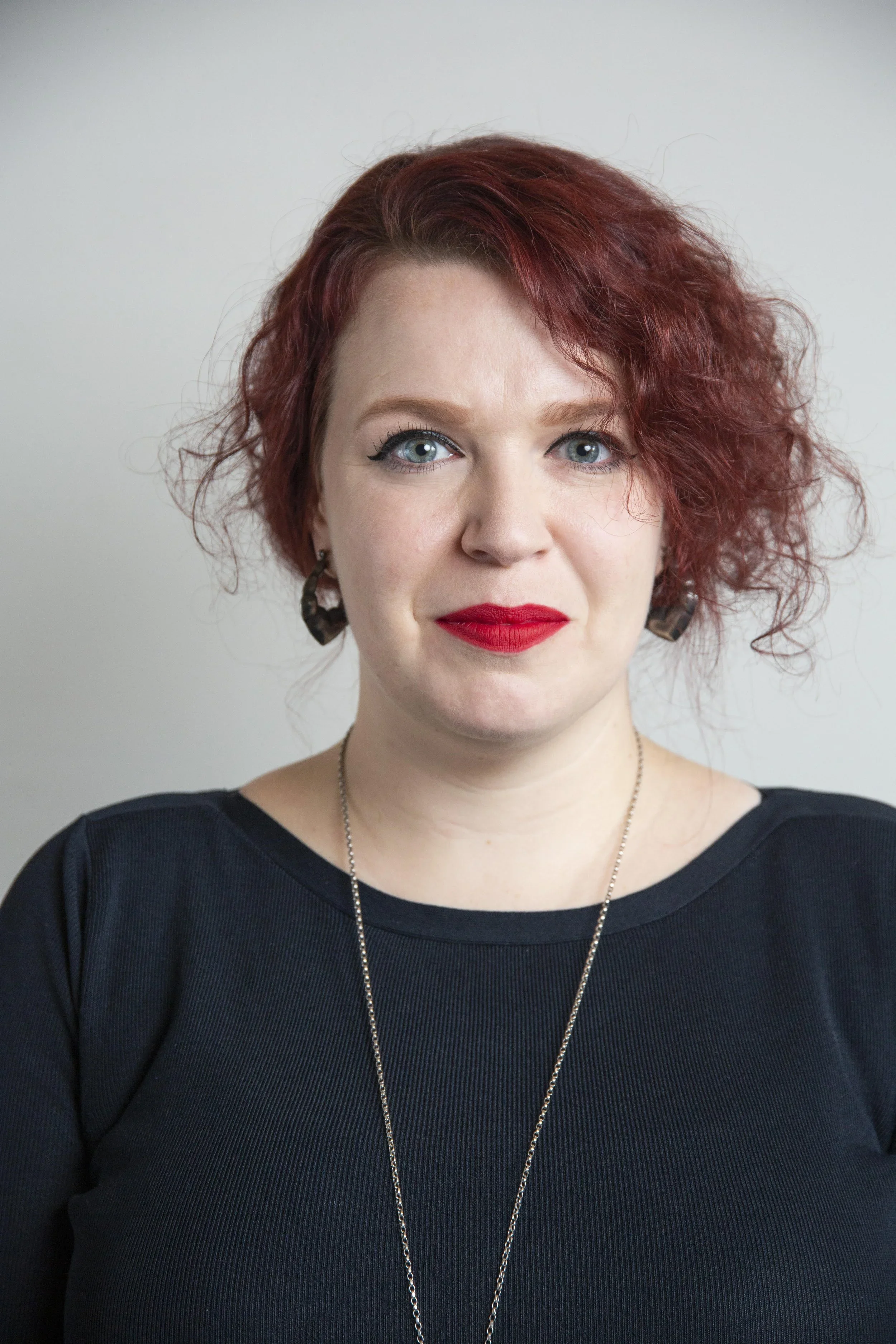 Close-up of a woman with red hair, blue eyes, red lipstick, wearing black earrings and a black top, against a plain light background.