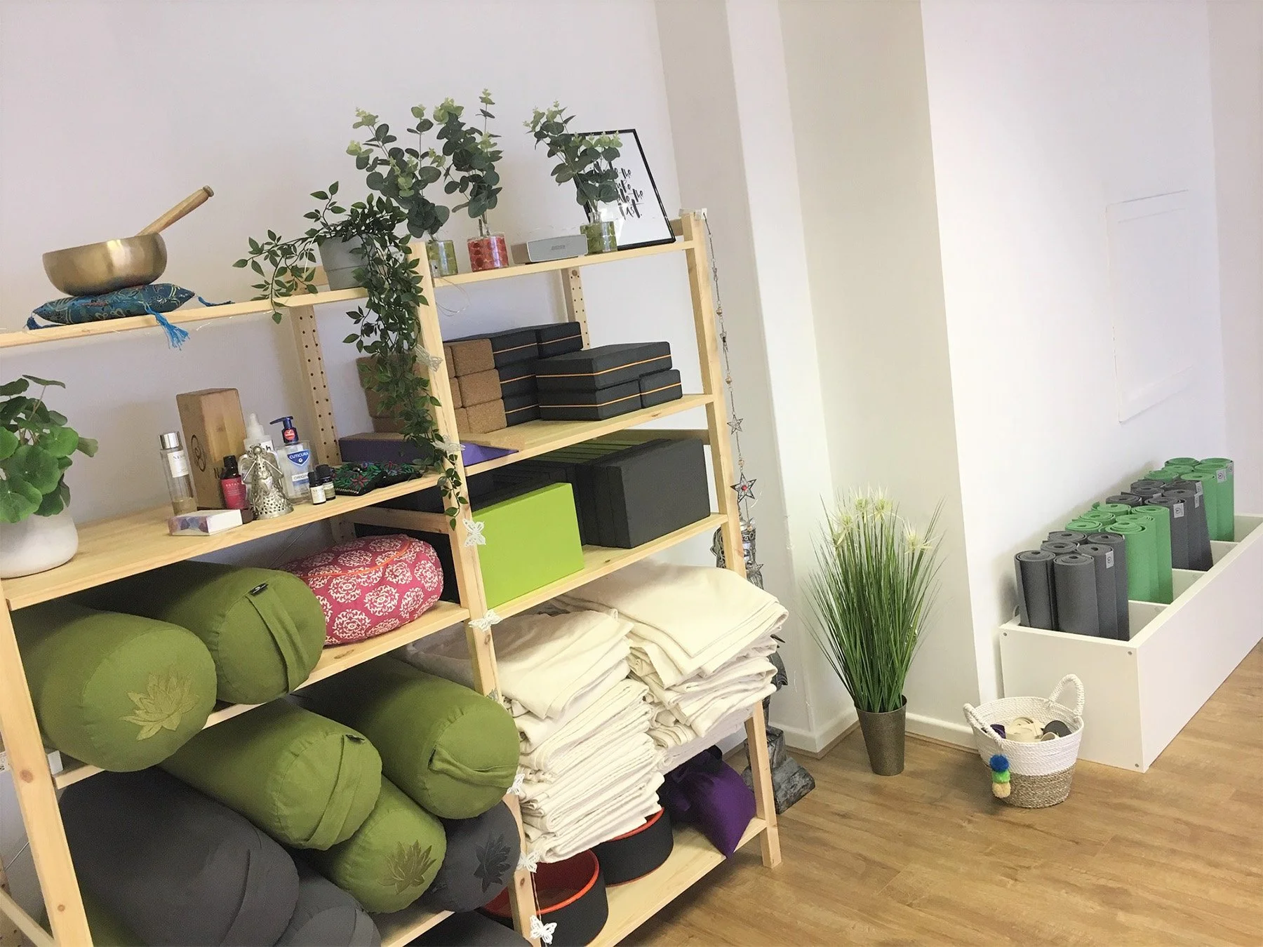 A wooden shelving unit in a wellness or yoga studio holds plants, cushions, folded blankets, and boxes. To the right, a white shelf contains rolled yoga mats in black and green, a potted plant with long grass, a basket with miscellaneous items, and a small pull toy. The room has wood flooring and white walls.