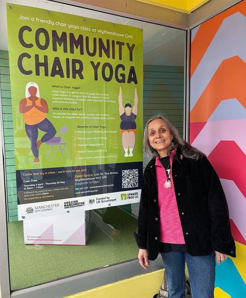 Woman standing next to a colorful poster advertising community chair yoga classes at Wythenshawe Civic, Manchester, UK.