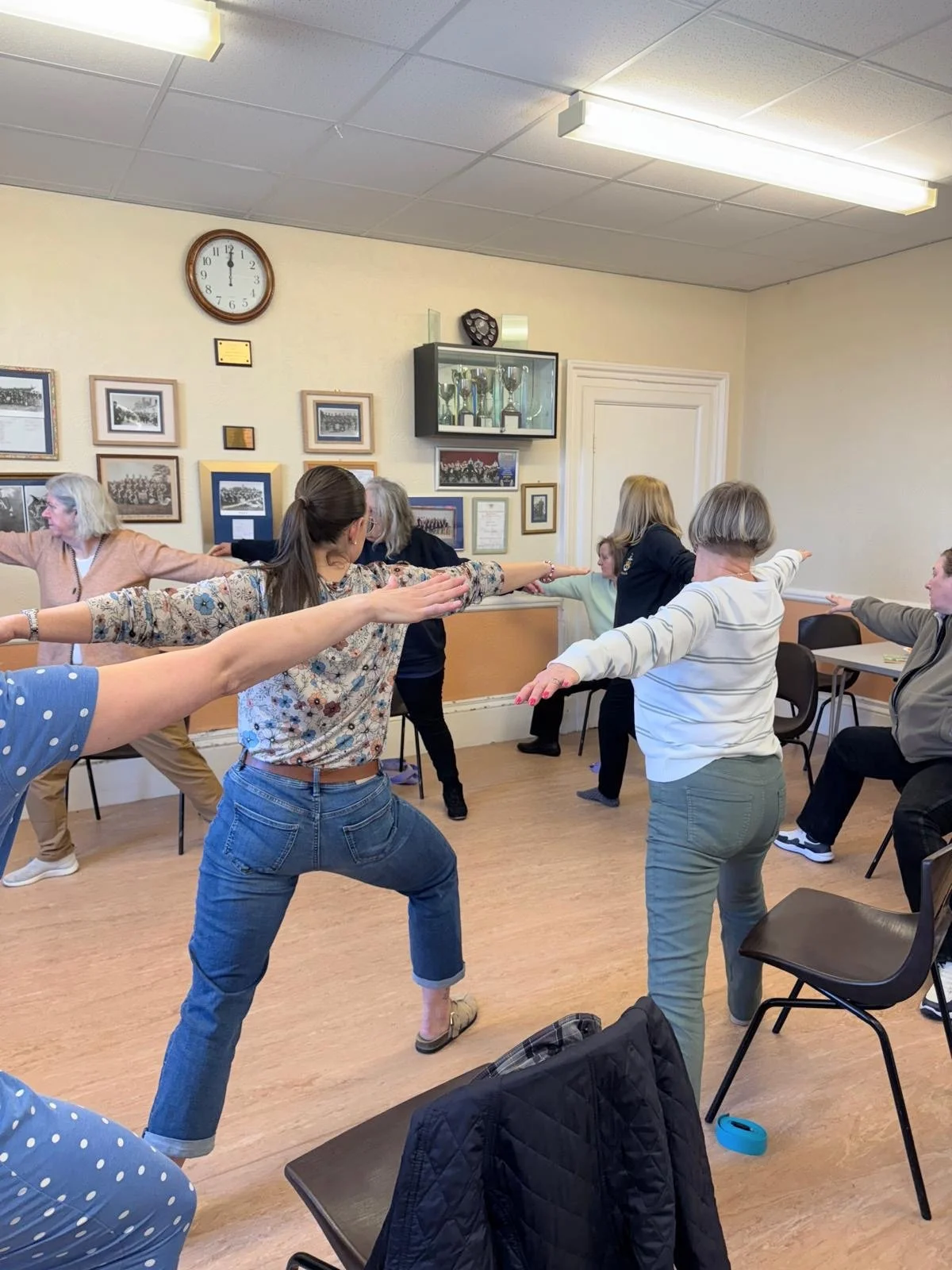 Group of women participating in a seated exercise class in a room with framed photos on the wall and a clock showing 12:00.