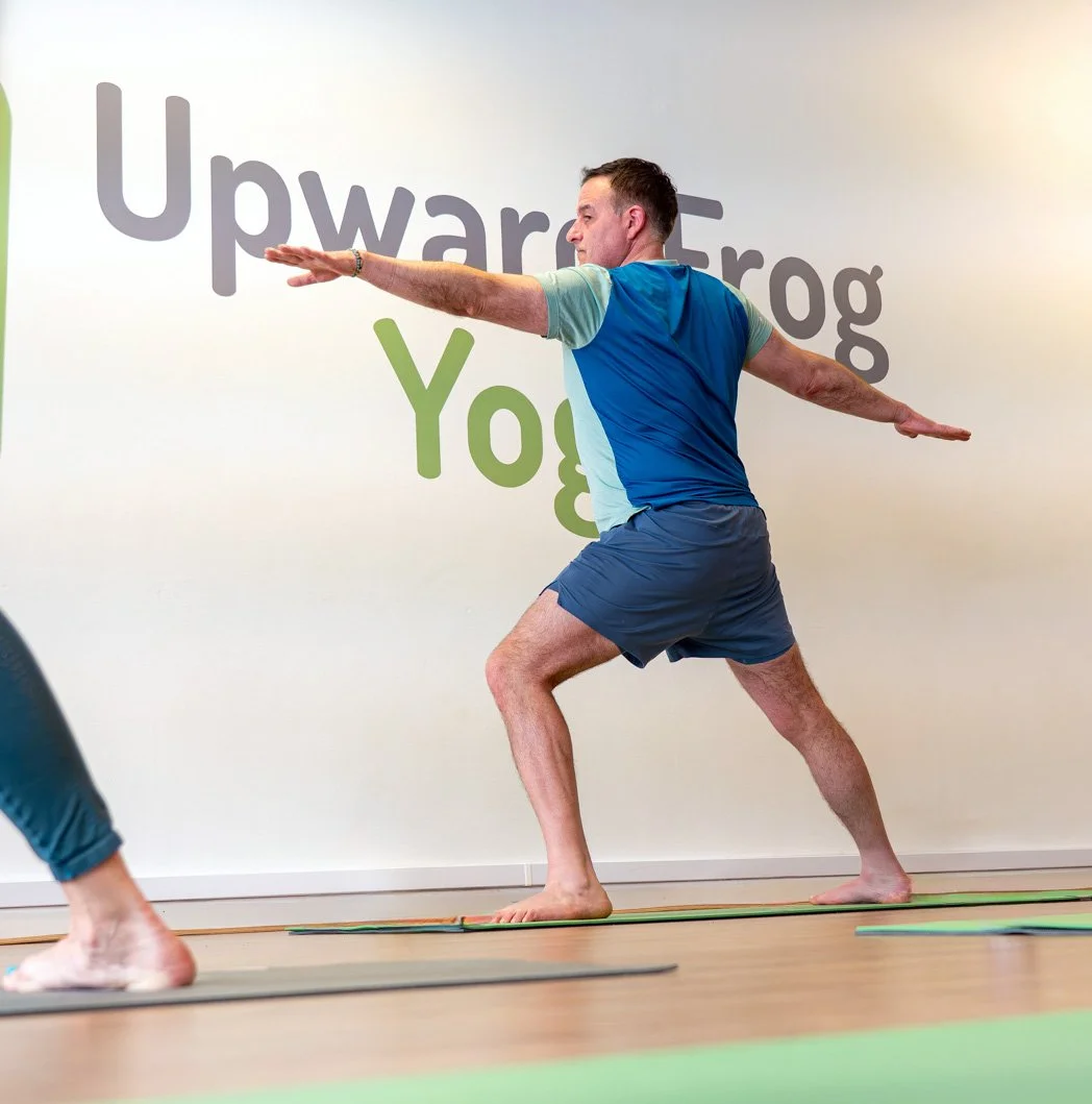 A man practicing yoga in a studio with a white wall in the background. The wall features large grey and green text reading "Upward Frog Yoga." The man is barefoot, wearing a blue and grey athletic shirt and navy shorts, and is in a Warrior pose, extending his arms outward and balancing on one foot.