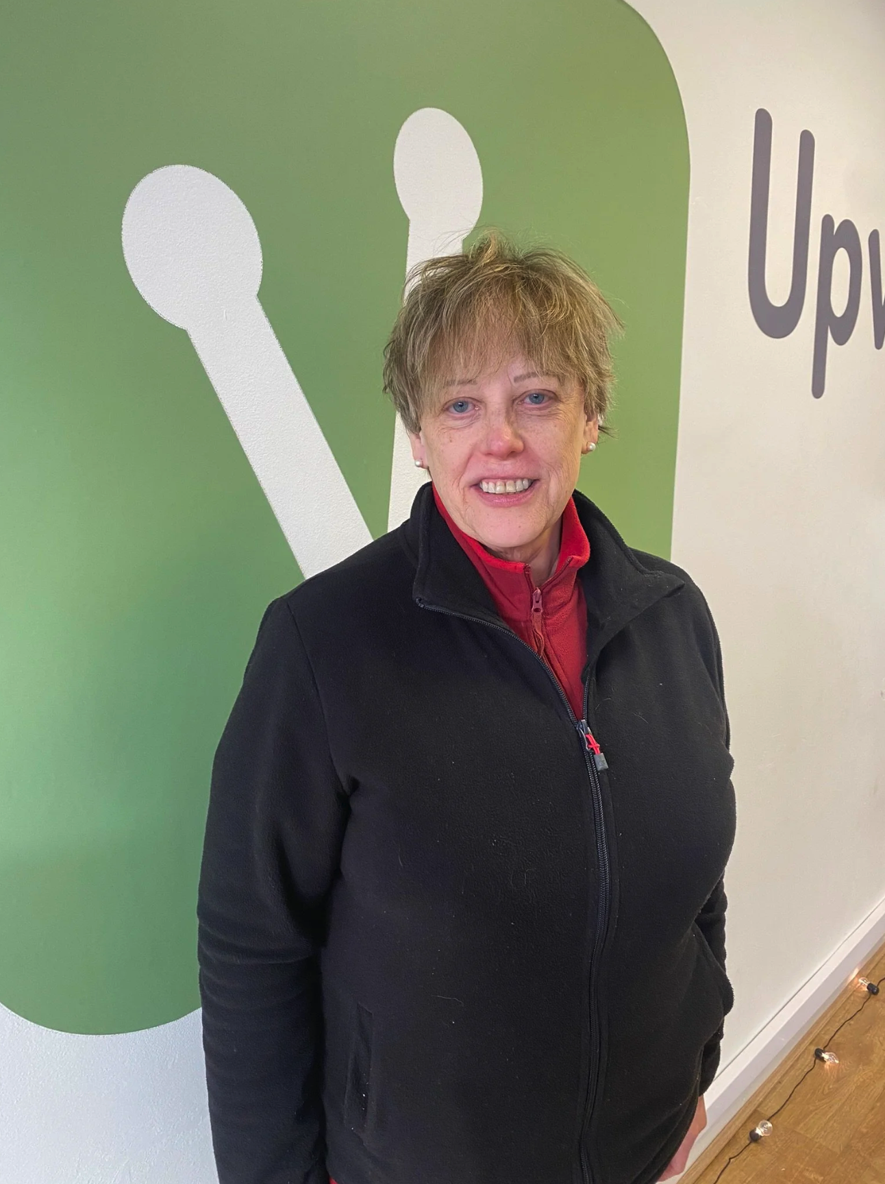 A woman with short, light brown hair and blue eyes standing indoors in front of a green and white wall with text and design. She is smiling and wearing a black zip-up jacket and a red shirt.