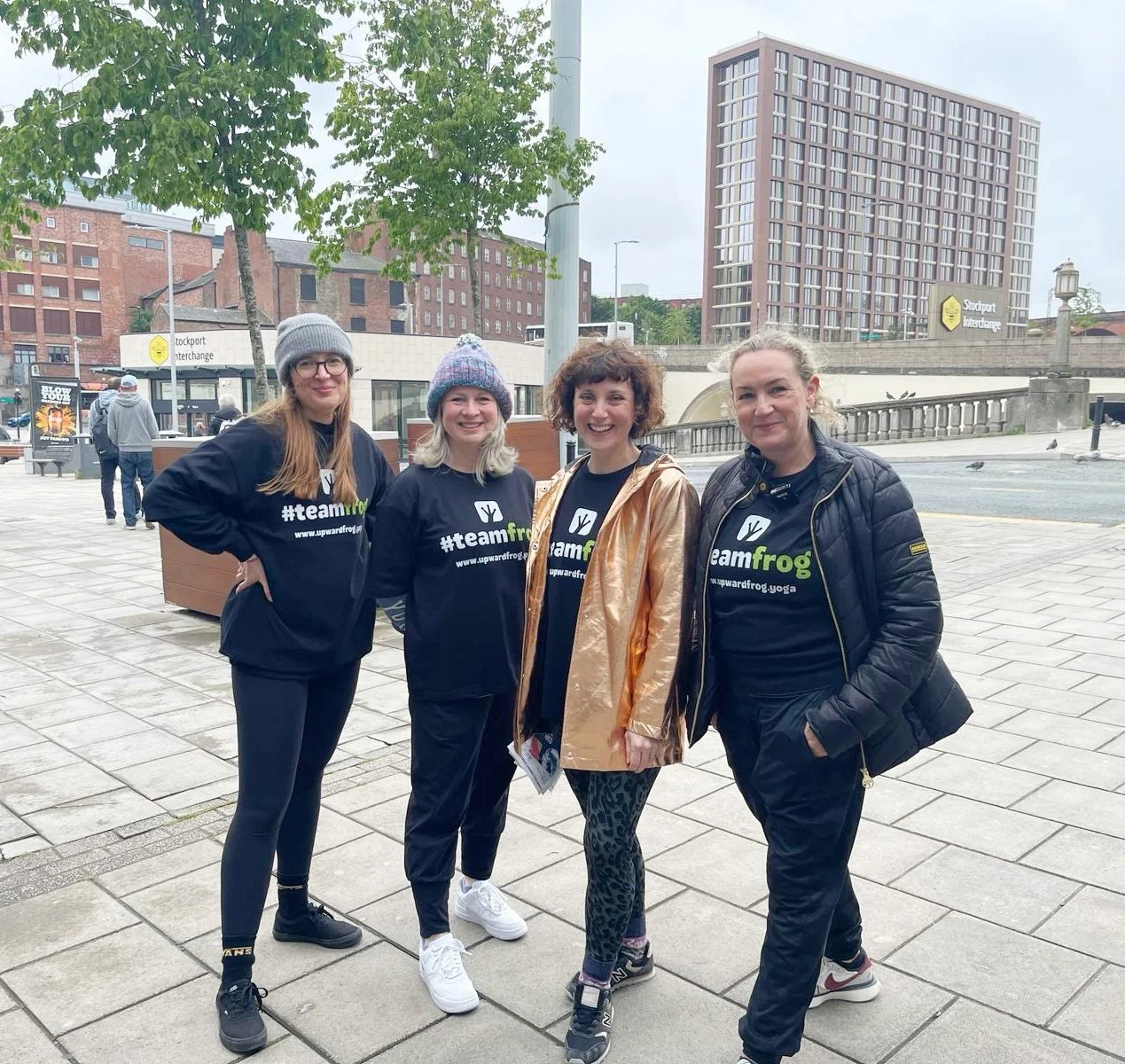 Four women standing outdoors in a city plaza, all wearing black t-shirts with the hashtag #teamfrog and a frog logo, smiling at the camera. Background includes buildings, trees, and a few pedestrians.