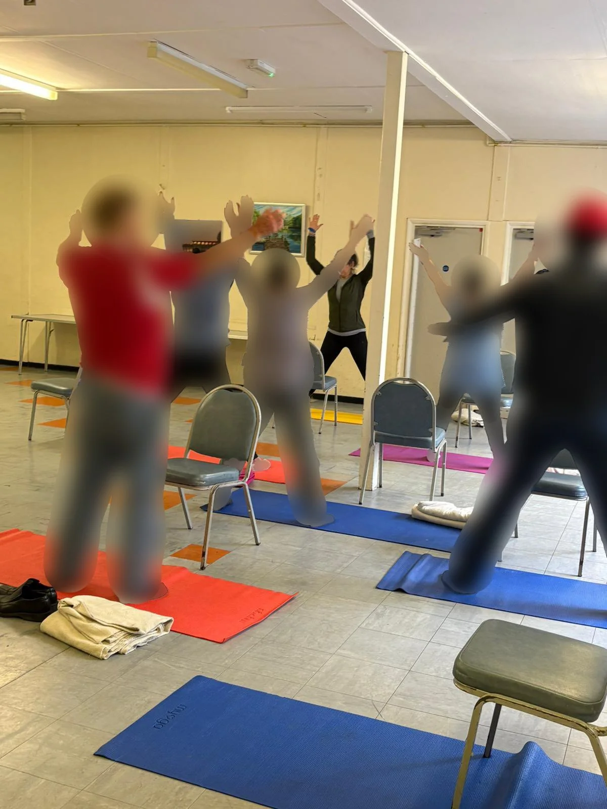 People participating in a seated yoga or stretching class in a room, with some individuals standing with arms raised, on colorful yoga mats.