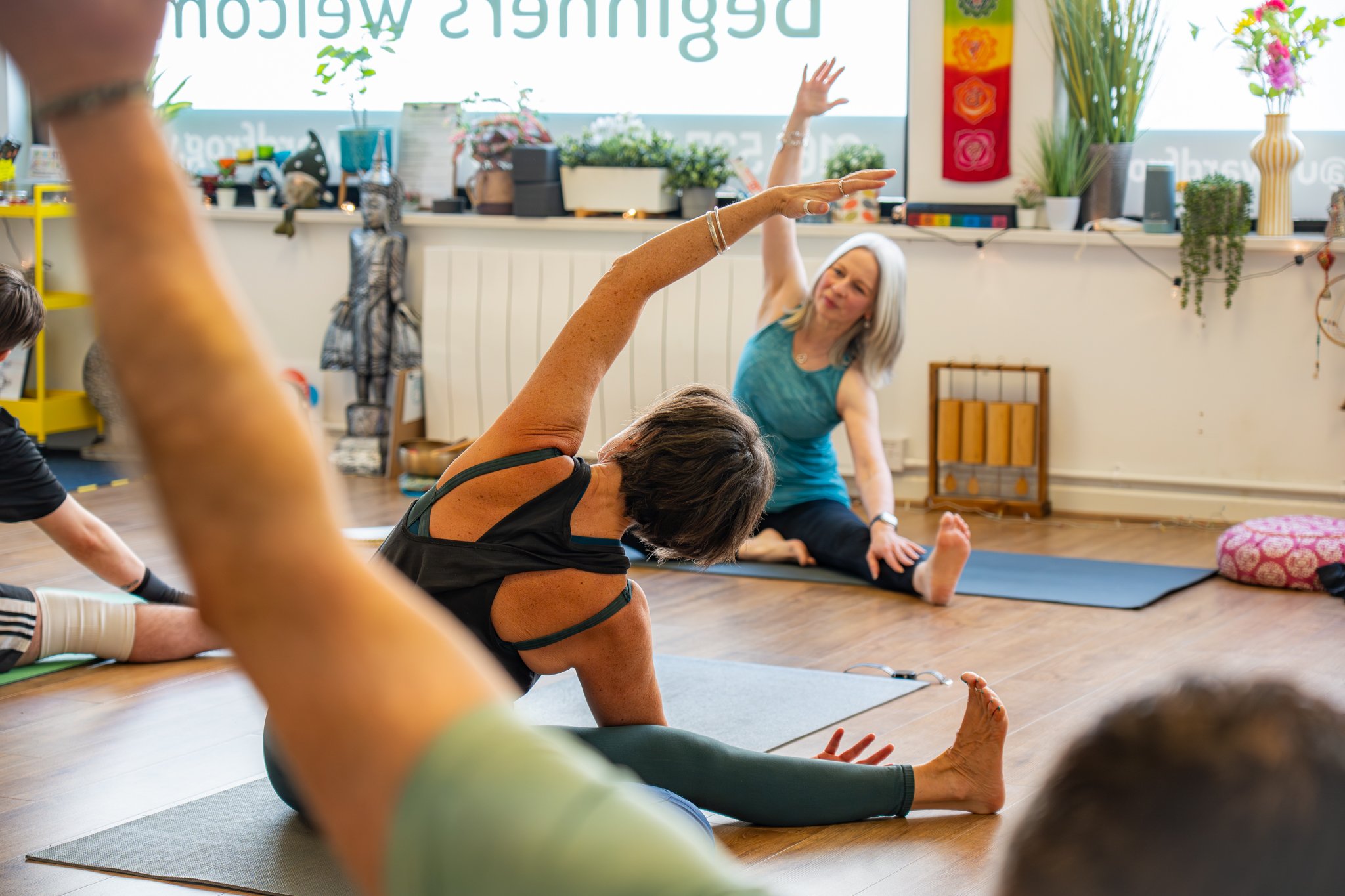 A group of people in a yoga class, practicing stretches and poses on yoga mats in a brightly decorated room with plants and colorful artwork.