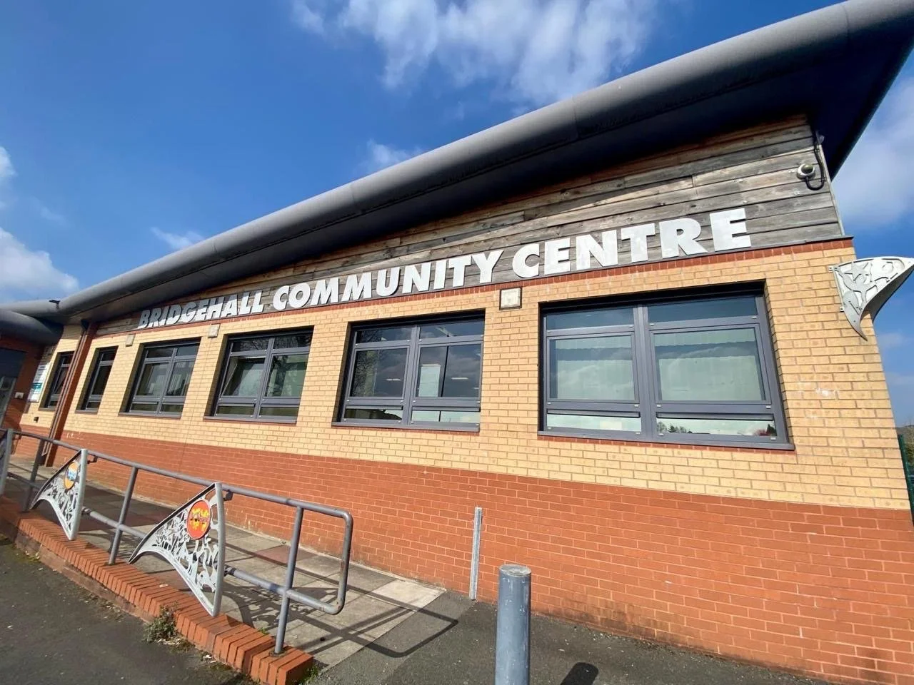 Exterior view of Bridge Hall Community Centre building with brick and wooden facade, large windows, metal railings, and decorative metal panels, under a partly cloudy sky.