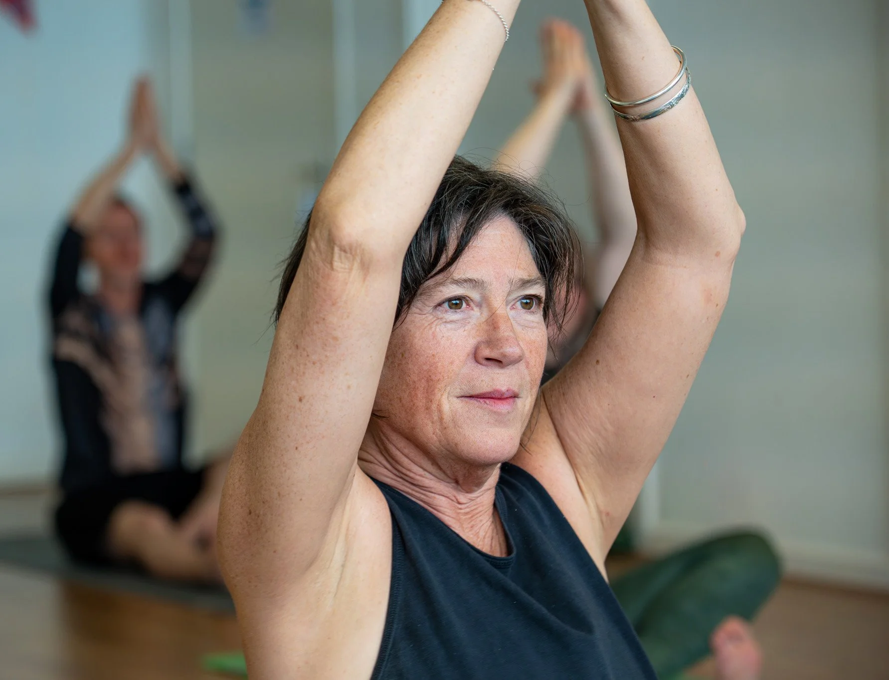 A woman with short dark hair and freckles performing a yoga pose with arms raised above her head in a yoga class, with other women practicing in the background.
