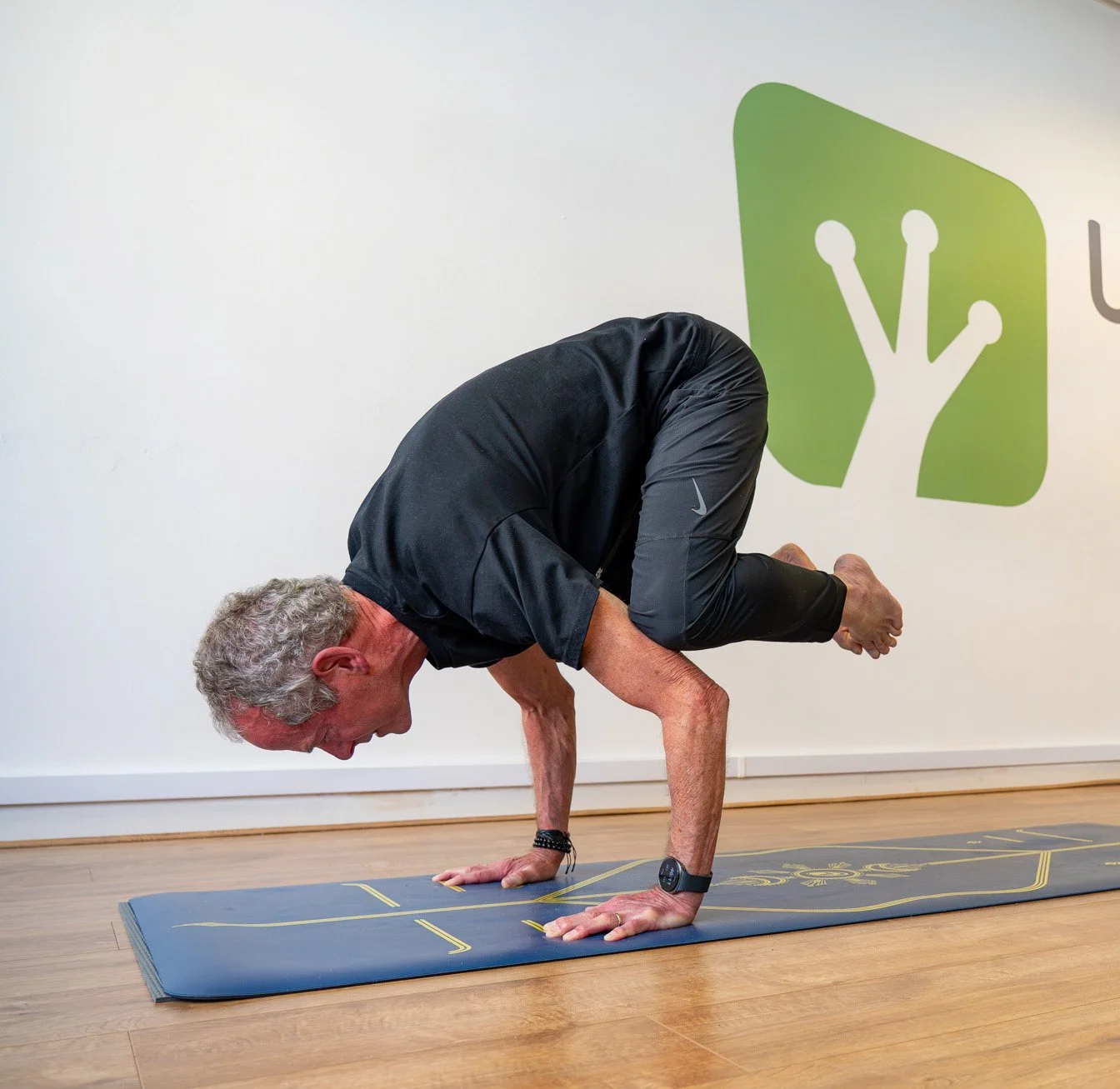 An elderly man performing a butterfly yoga pose on a blue mat in a fitness studio with a green logo on the wall.