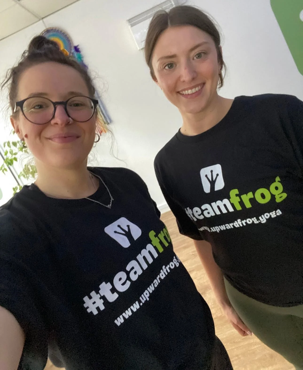 Two women wearing black t-shirts with the hashtag #teamfrog and a yoga pose logo. They are smiling and taking a selfie indoors.