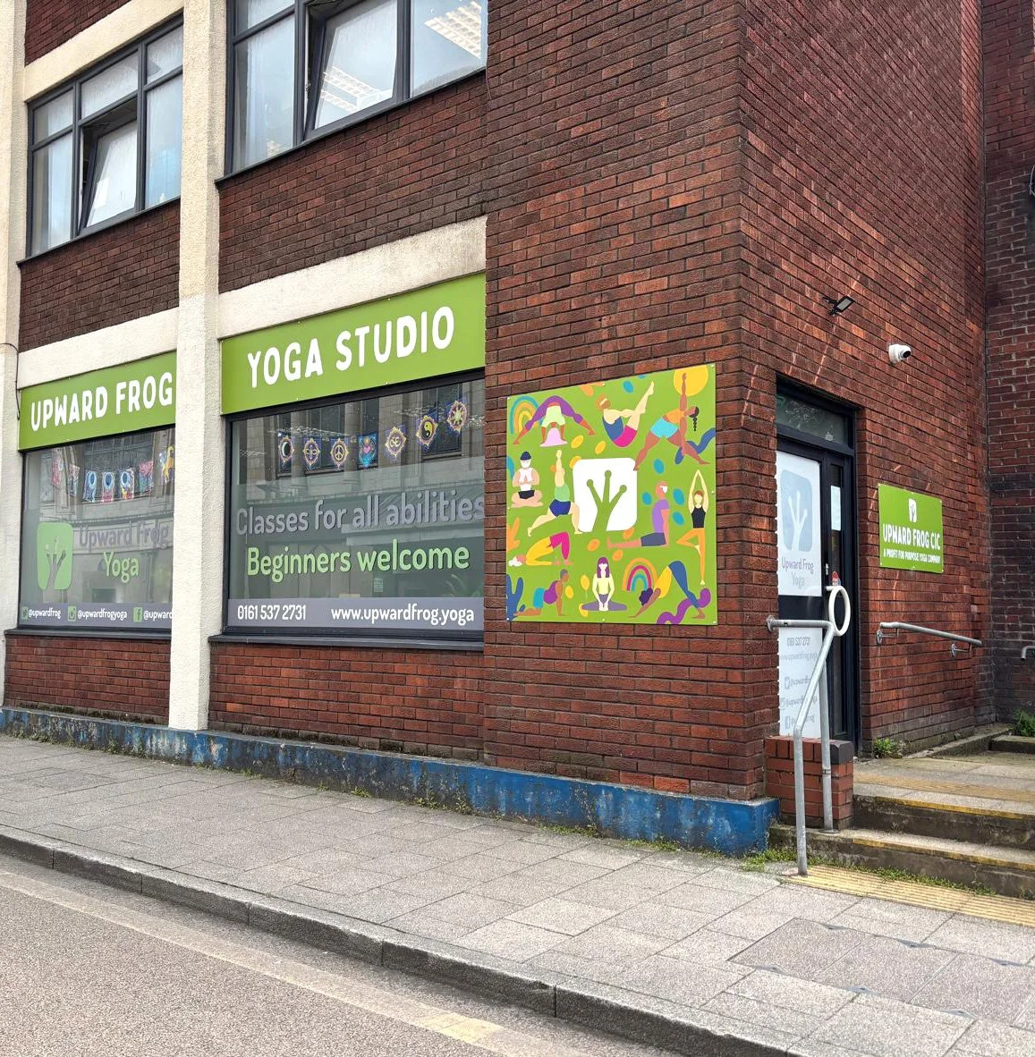 The exterior of a brick building with green signs for "Upward Frog Yoga Studio" displaying classes for all abilities and beginners welcome. There are colorful illustrations of people practicing yoga on the wall outside.