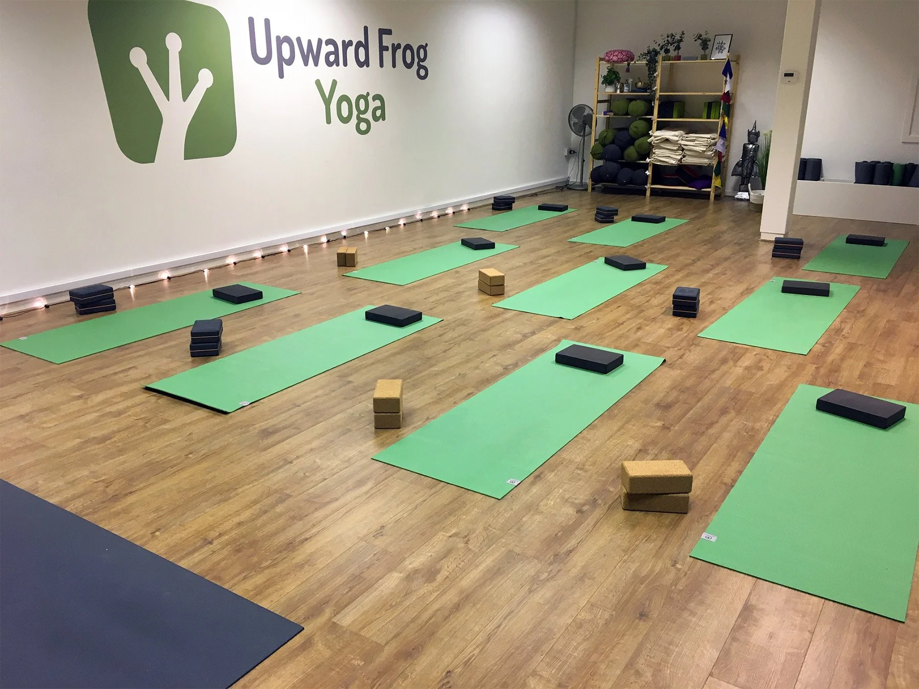 A yoga studio with green mats and black yoga blocks arranged on a wooden floor. The wall has a logo for Upward Frog Yoga. In the background, shelves hold yoga props like blankets, bolster cushions, and plants, with some decorative items and a fan nearby.