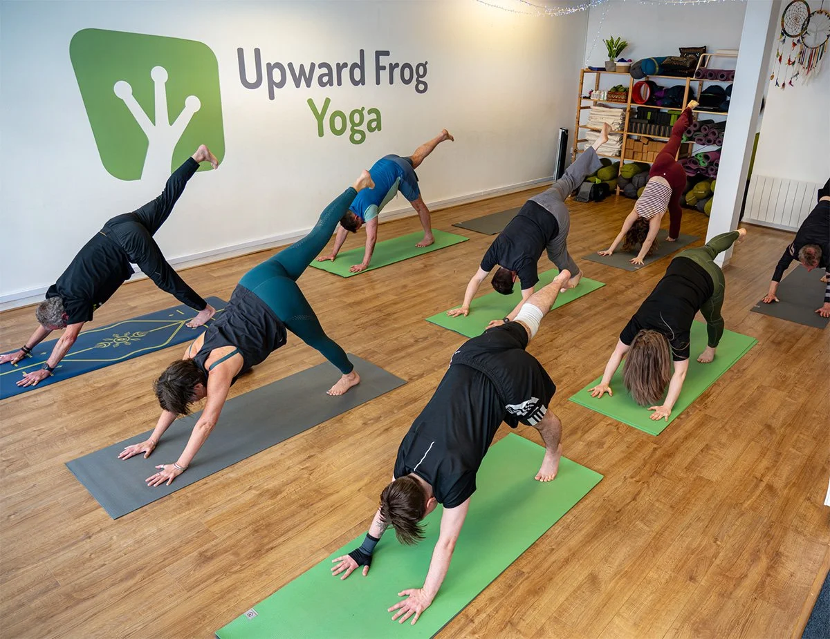 People practicing yoga in a studio with wooden flooring, mats, and a wall sign reading 'Upward Frog Yoga' with a logo.