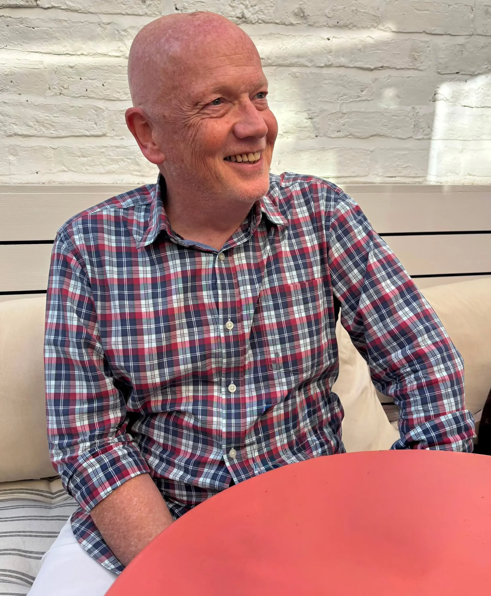 An older man with a bald head and wearing a red, blue, and white plaid shirt, sitting at a table with a red surface, smiling and looking to his left in an indoor setting.