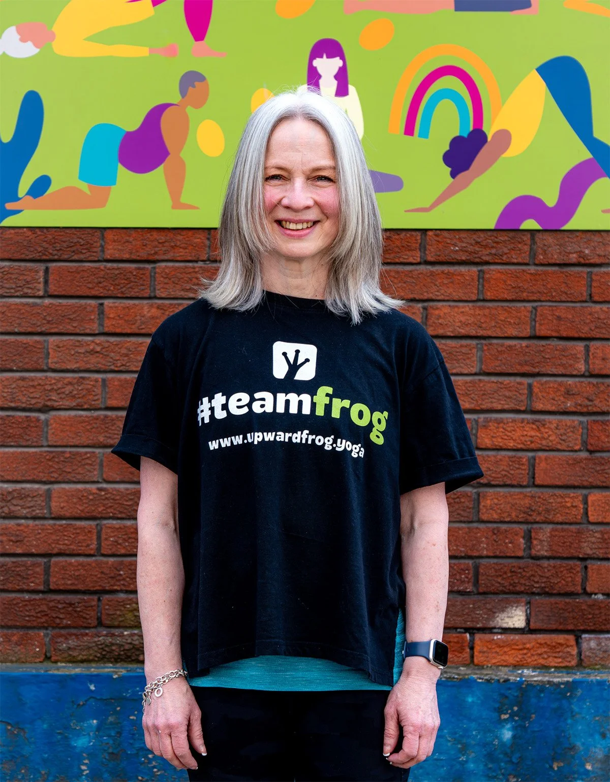 A woman standing in front of a brick wall with a colorful mural of people and rainbows. She is smiling and wearing a black t-shirt with the hashtag #teamfrog and a website URL.