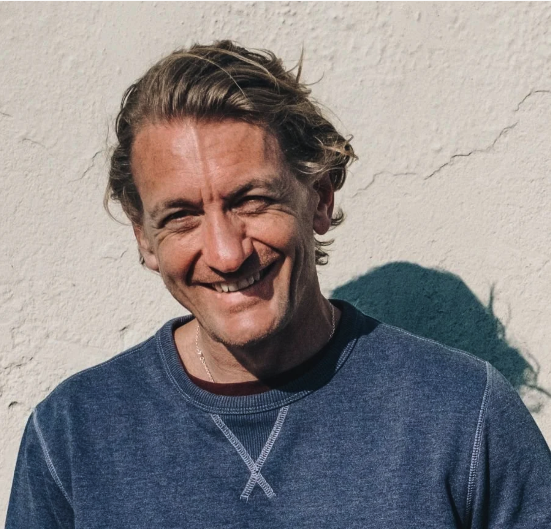 A man smiling, with wavy blonde hair, wearing a navy blue shirt, standing against a textured white wall.