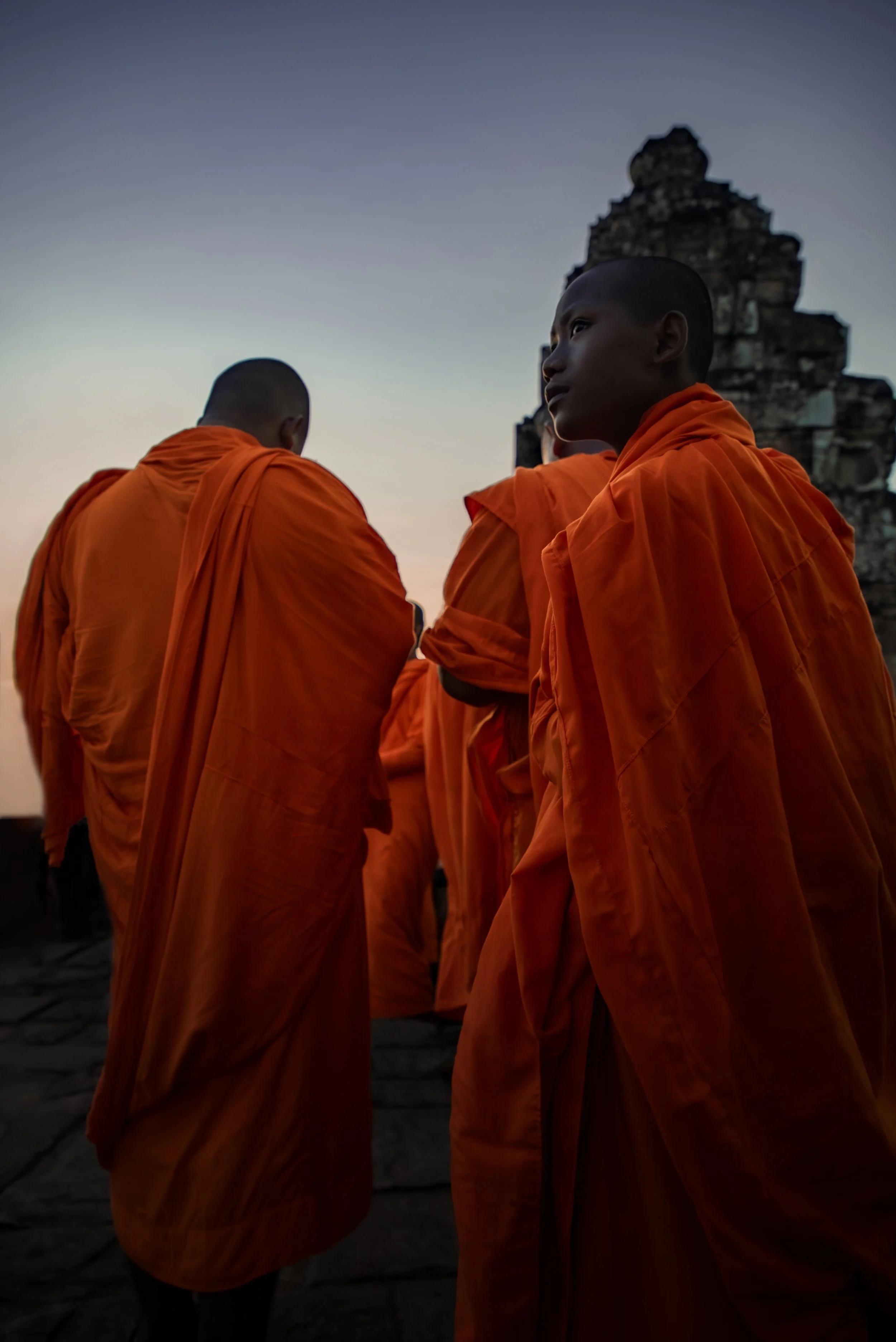 Monks wearing orange robes standing outdoors during sunset, with an ancient stone structure in the background.