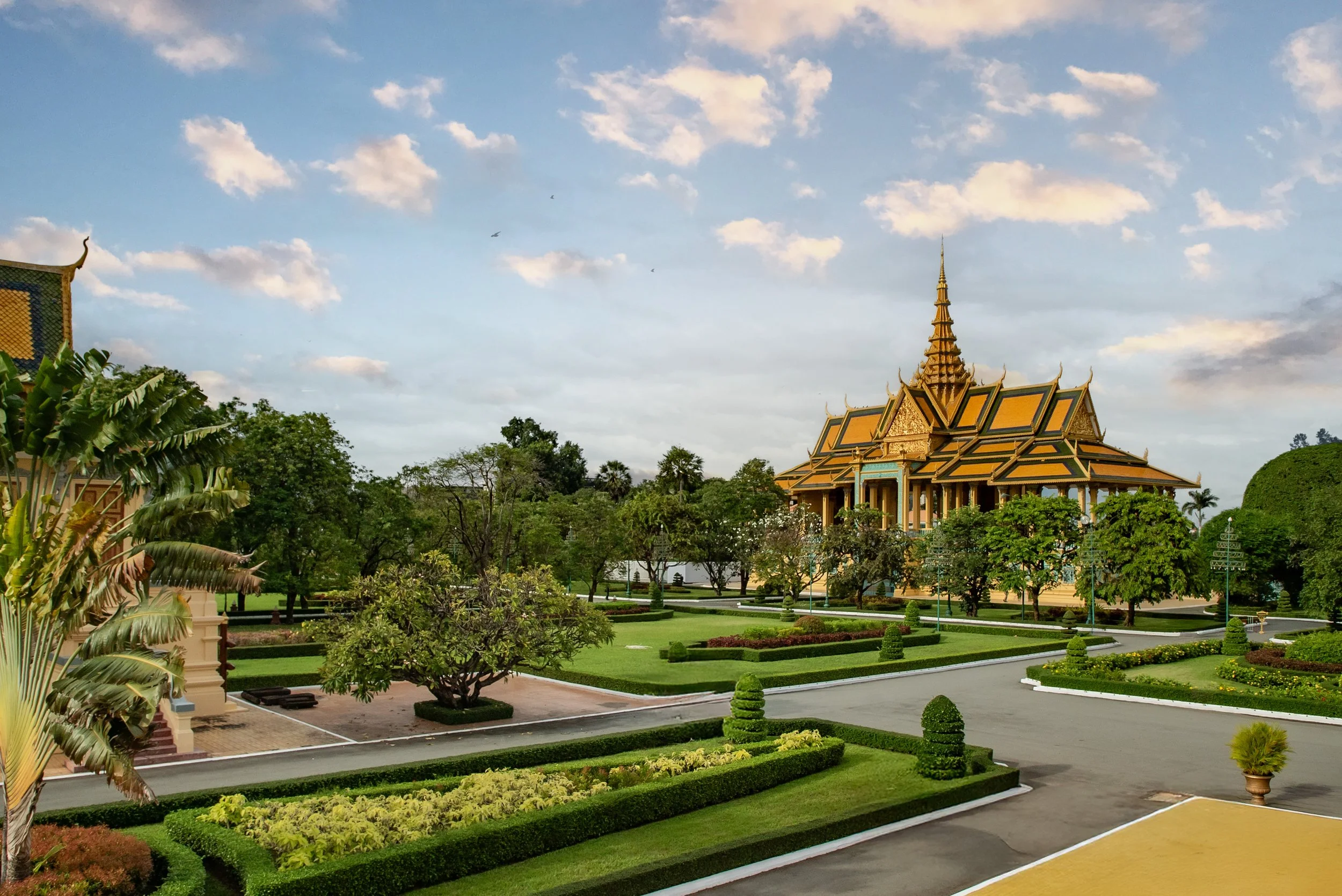 Traditional Thai temple with ornate golden roof, surrounded by lush greenery and manicured gardens under a partly cloudy sky.