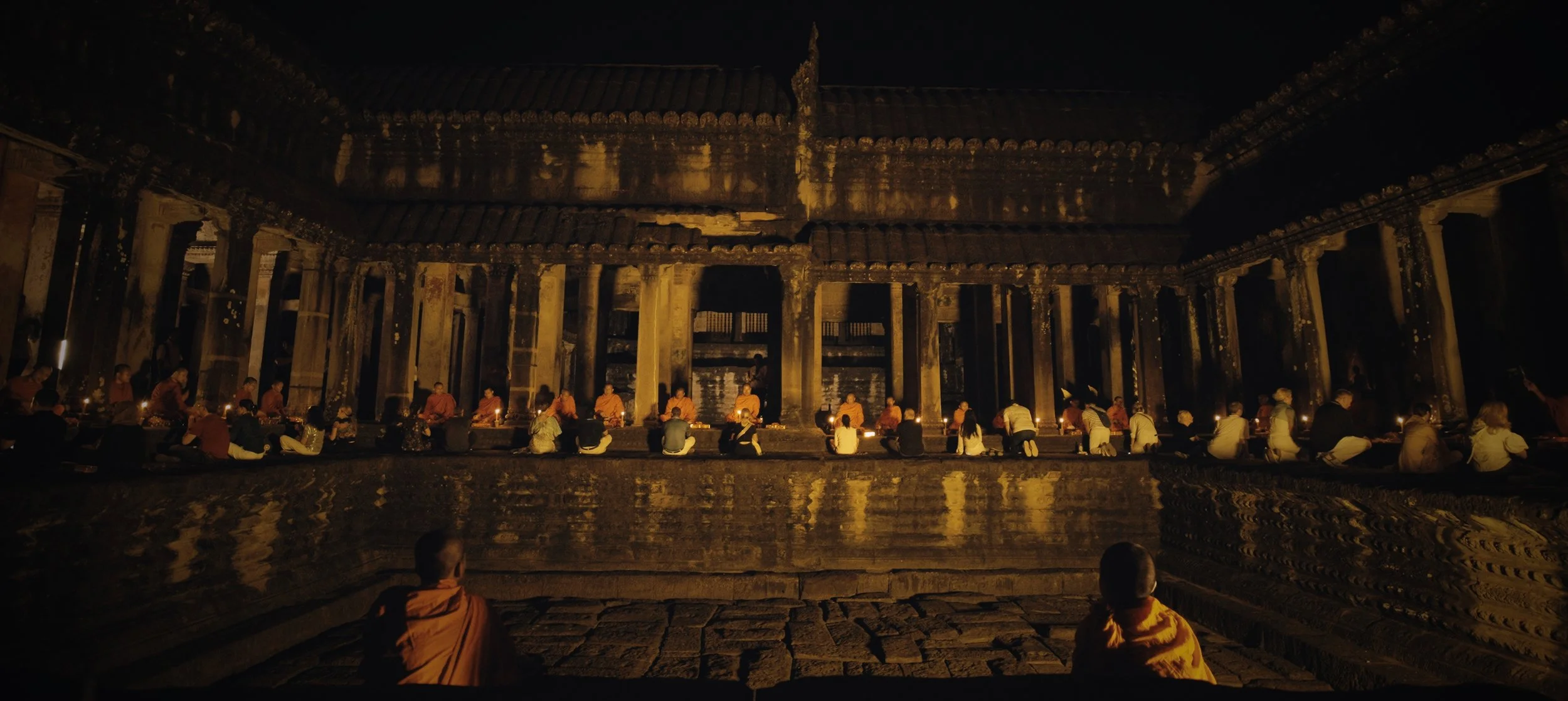 Monks and laypeople gather at night inside a historic temple, engaged in a candlelit ceremony with traditional attire and dim lighting.