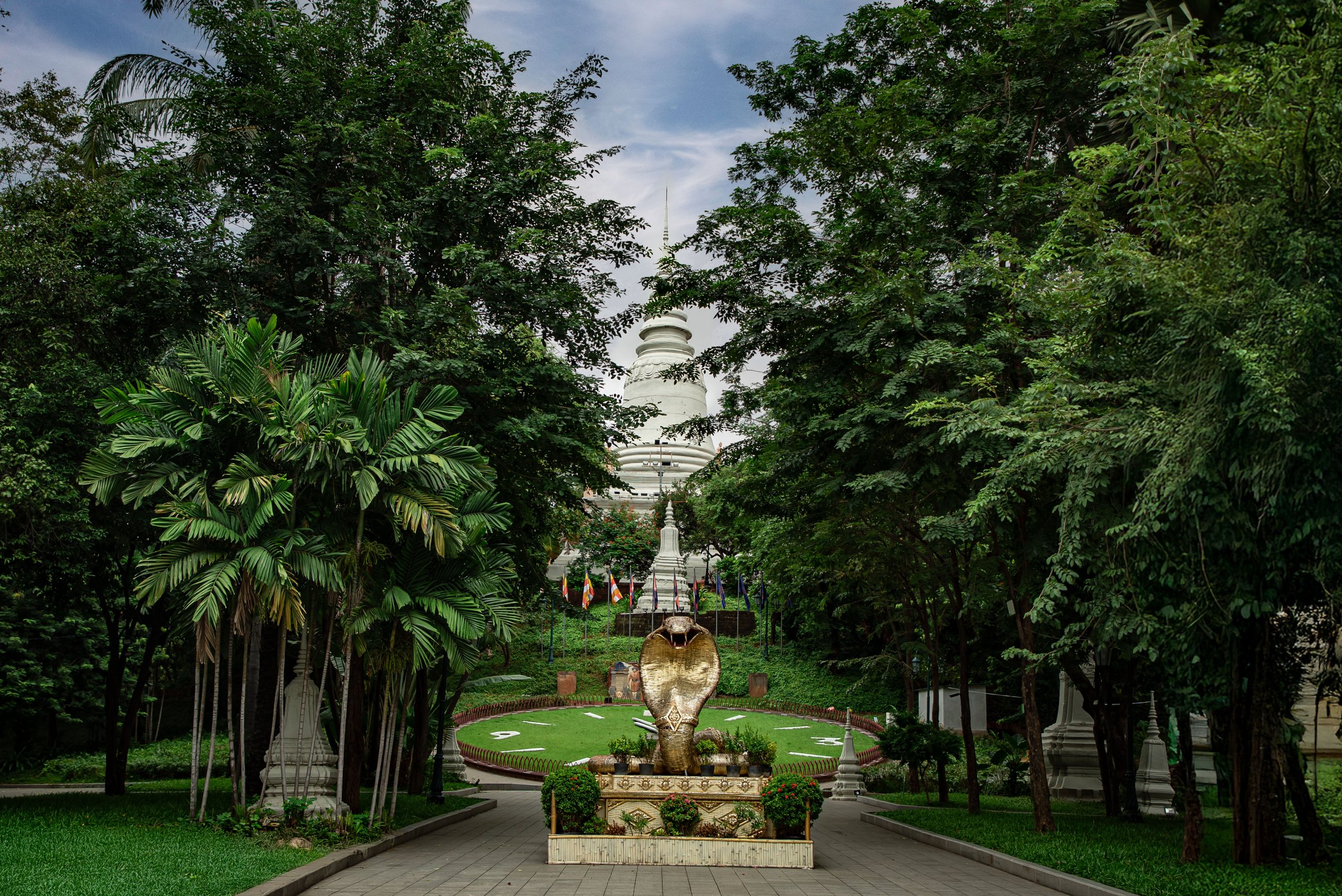 A garden pathway leads to a white stupa in the distance, with a large elephant head sculpture in the foreground, surrounded by lush green trees and plants.