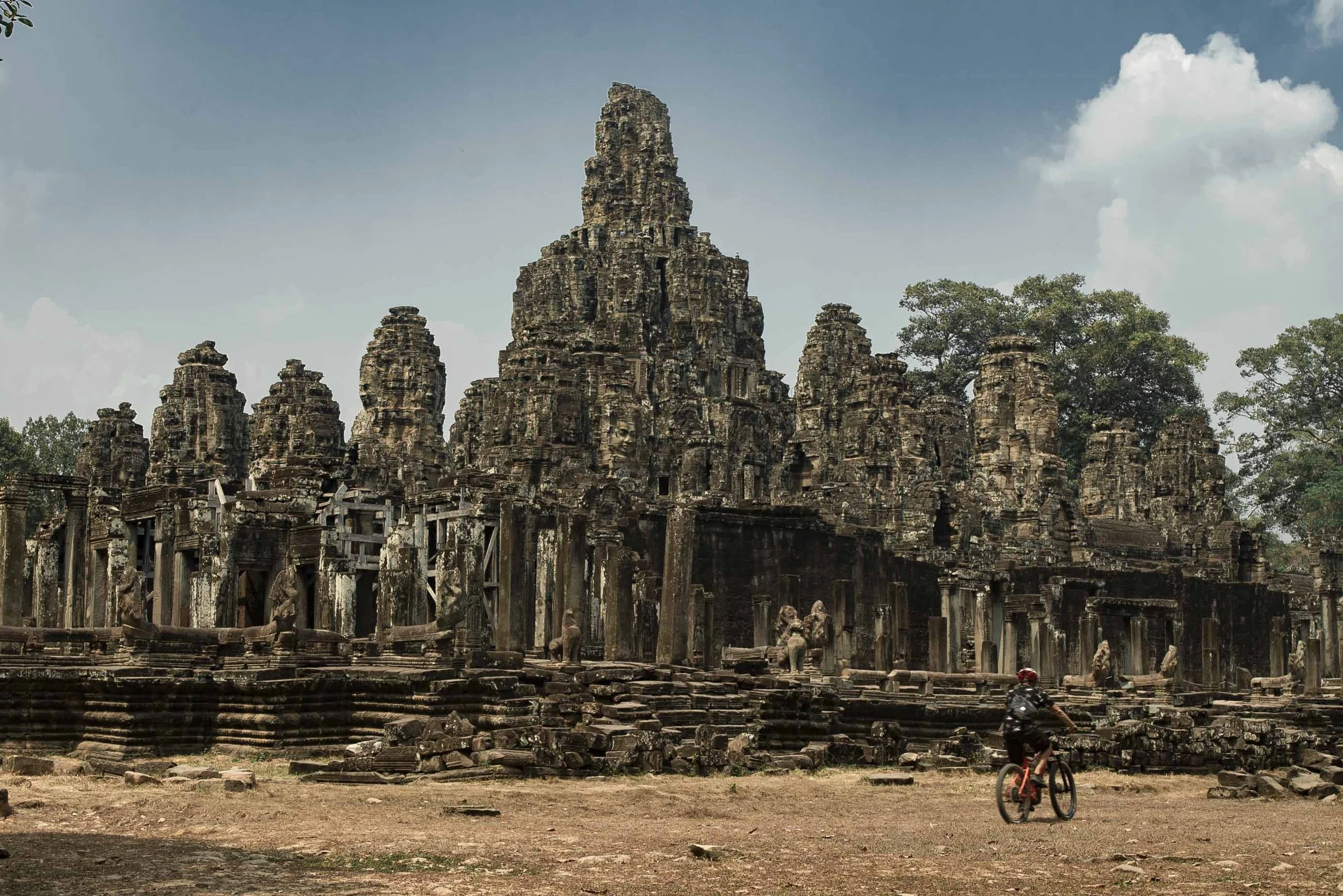 An ancient stone temple with carved faces and towers, with a cyclist riding in the foreground and trees in the background