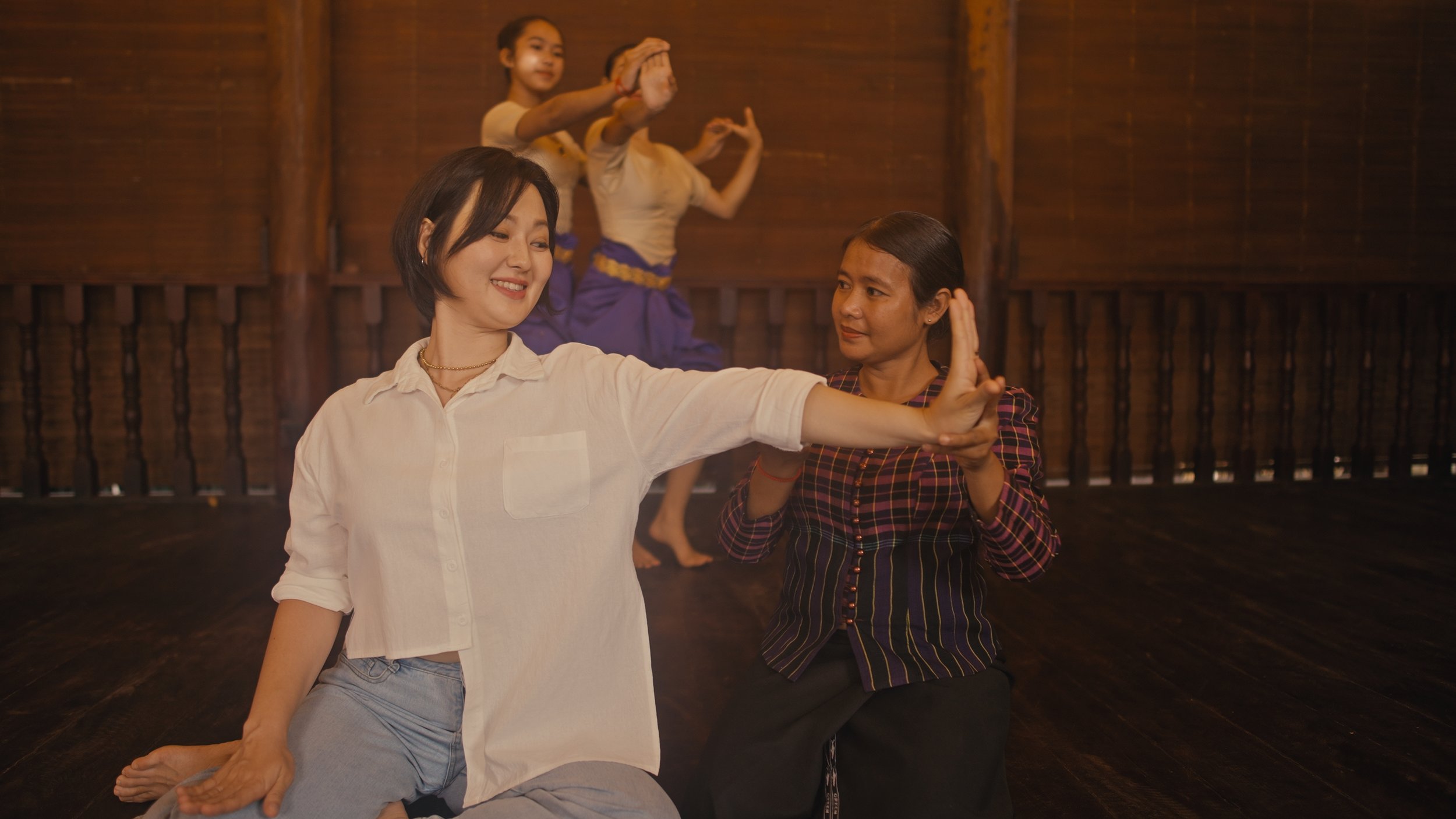 Two women practicing a traditional dance in a wooden hall while two girls perform a dance in the background.