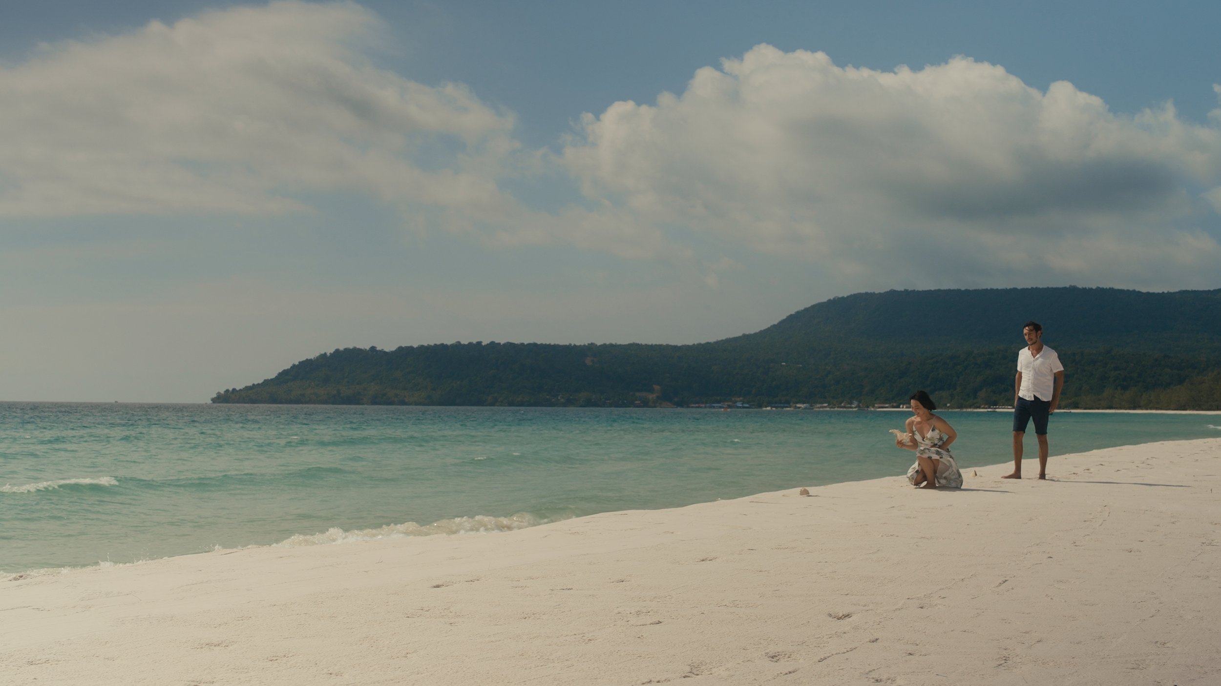 A woman kneeling on the beach holding a seashell and a man standing nearby on the sand with a mountain view in the background under a partly cloudy sky.
