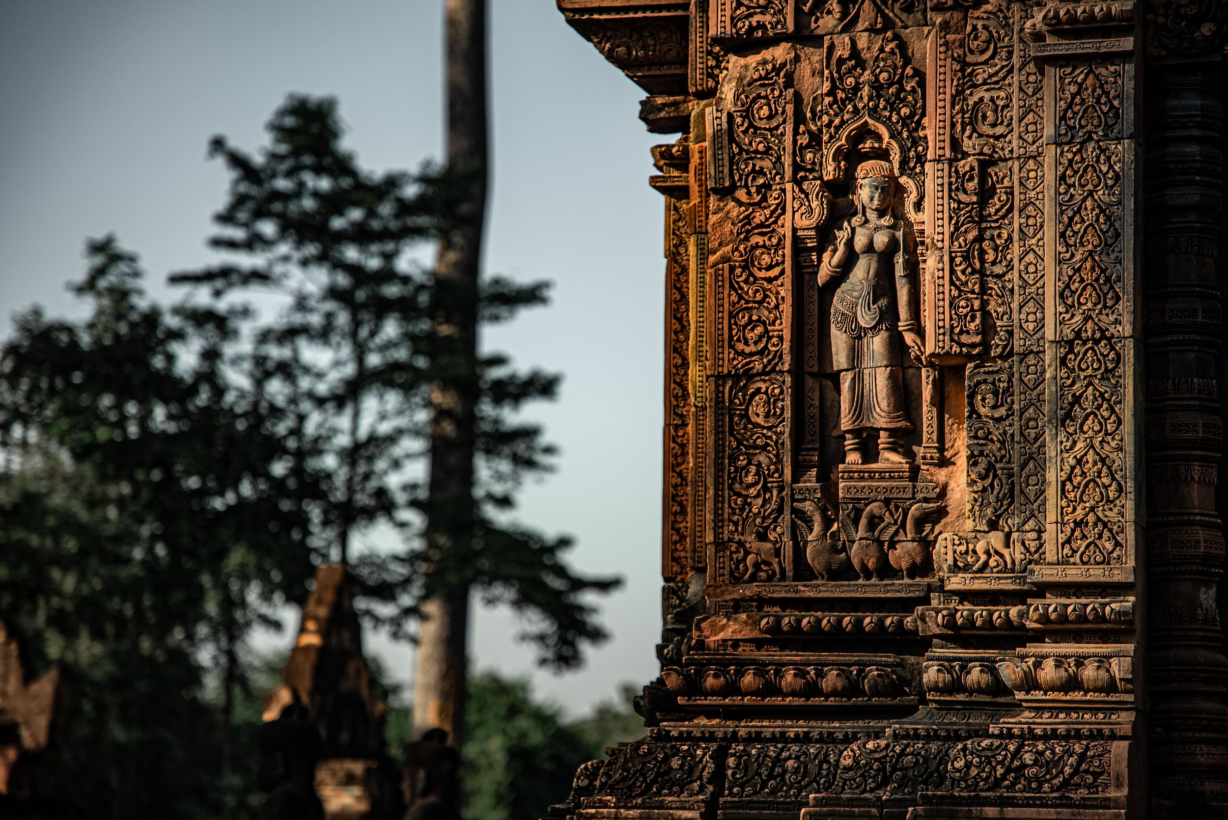 Intricate stone carvings of a standing female deity on a temple wall with lush green trees in the background.