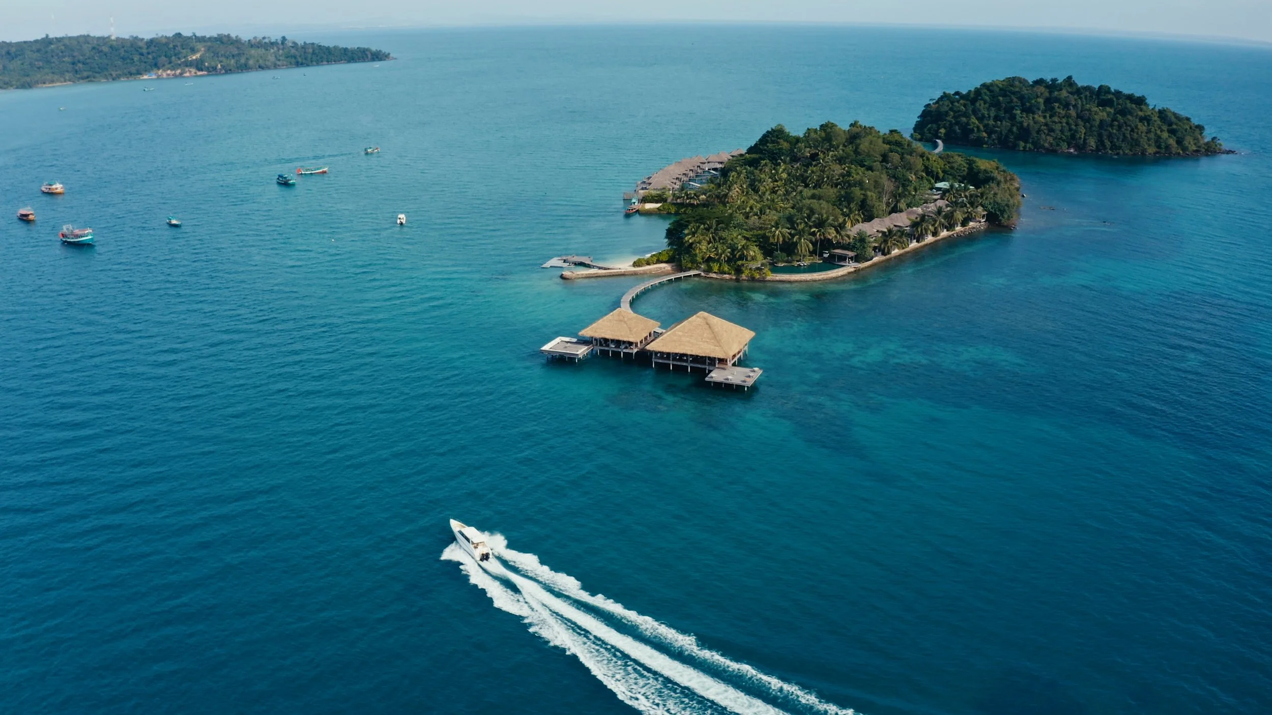 Aerial view of a tropical island resort with overwater bungalows, surrounded by turquoise water, boats, and lush green islands in the background.