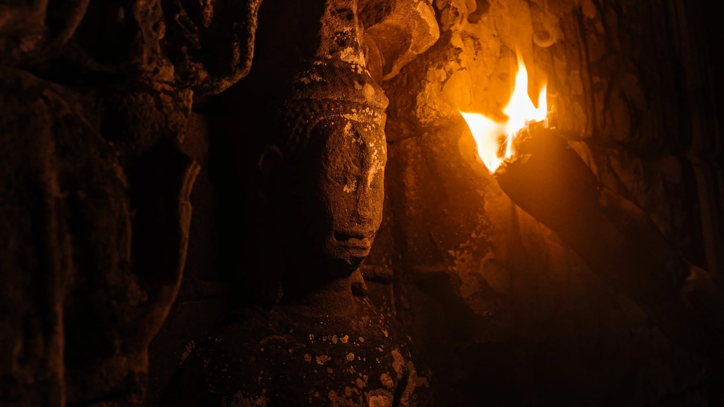 A stone Buddha statue illuminated by a torch with a flickering flame, set against a dark background.