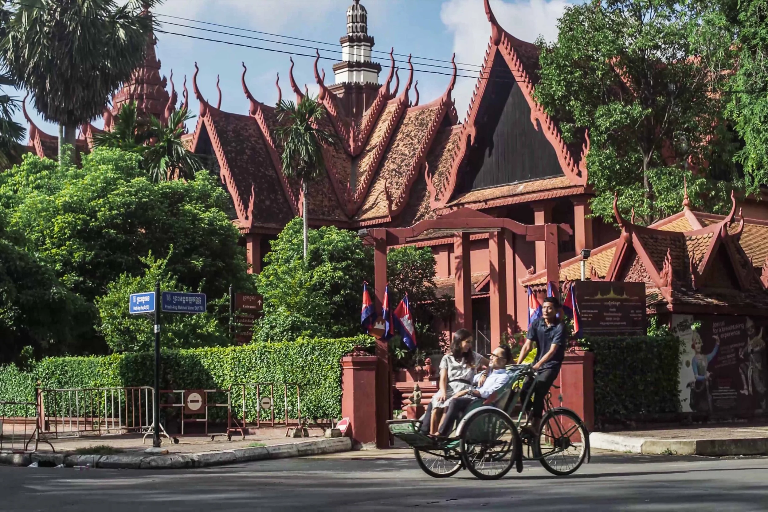Traditional Cambodian temple with intricate roofing and flags, two women and a man in a wheelchair being pushed on a bicycle, green trees and avenue signs in the background.